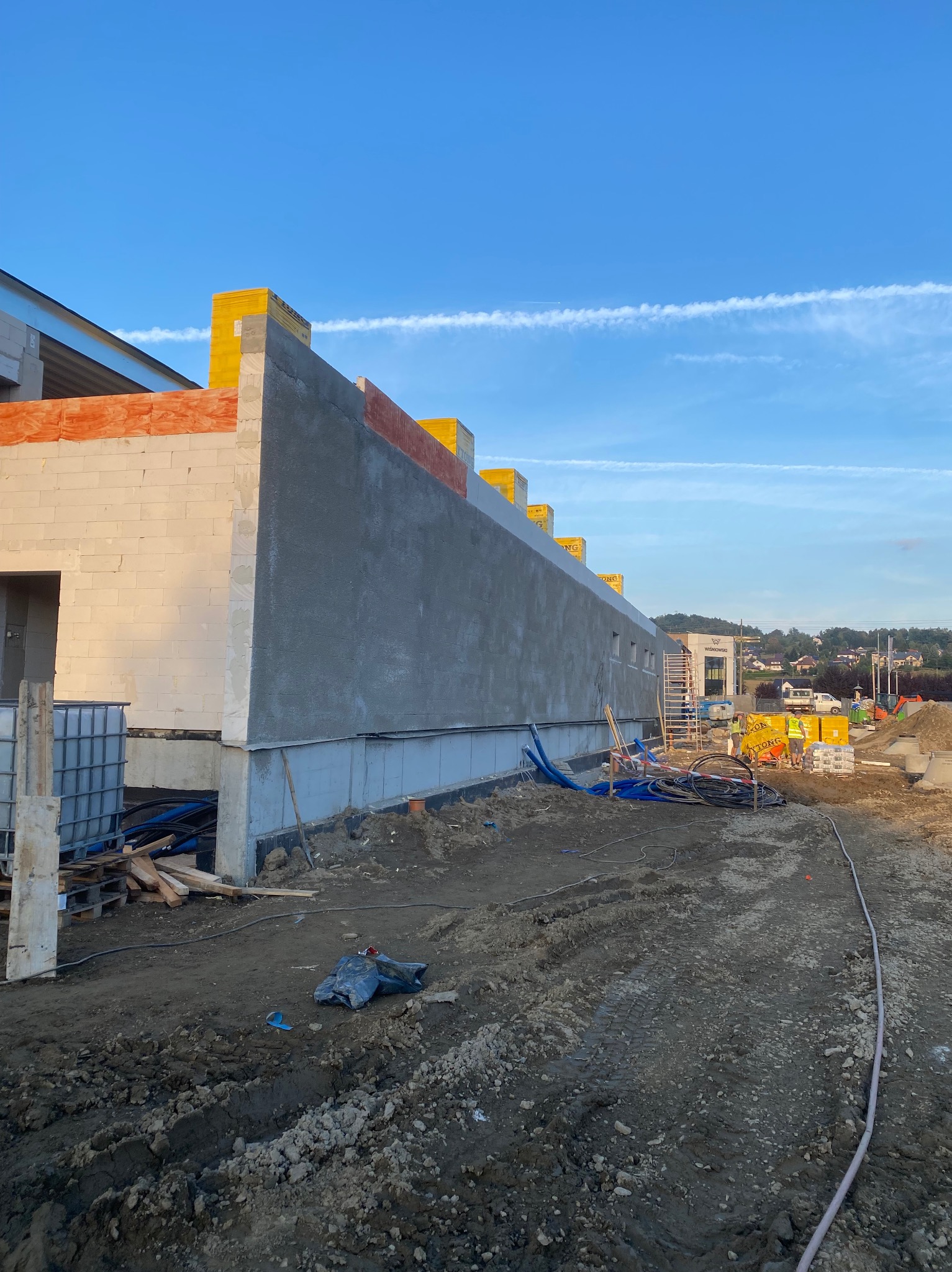 Ściana budynku w trakcie szpachlowania z widocznymi blokami Ytong, żółtą wełną mineralną na górze i świeżo nałożoną szarą szpachlą, niebieskie rury na ziemi, w tle budynki i wzgórza pod błękitnym...