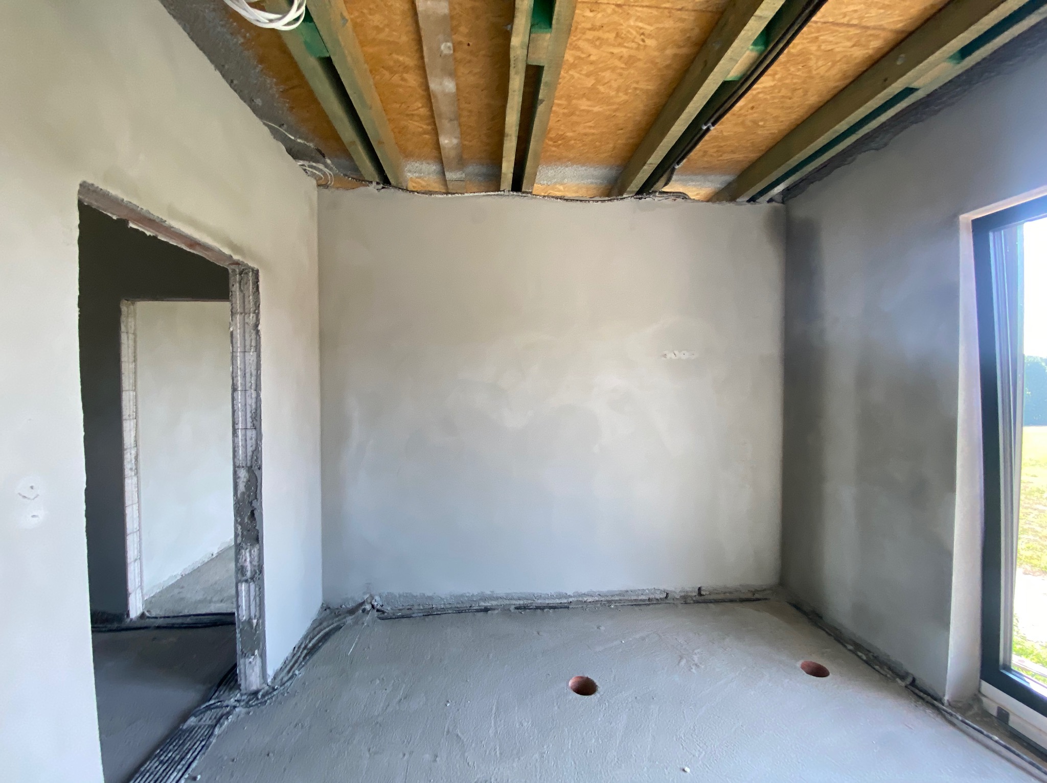 Surowe wnętrze w trakcie prac wykończeniowych: ściany pokryte świeżo nałożoną warstwą szpachli, widoczne przewody elektryczne przy podłodze i pod sufitem, sufit z drewnianą konstrukcją, wylewka...