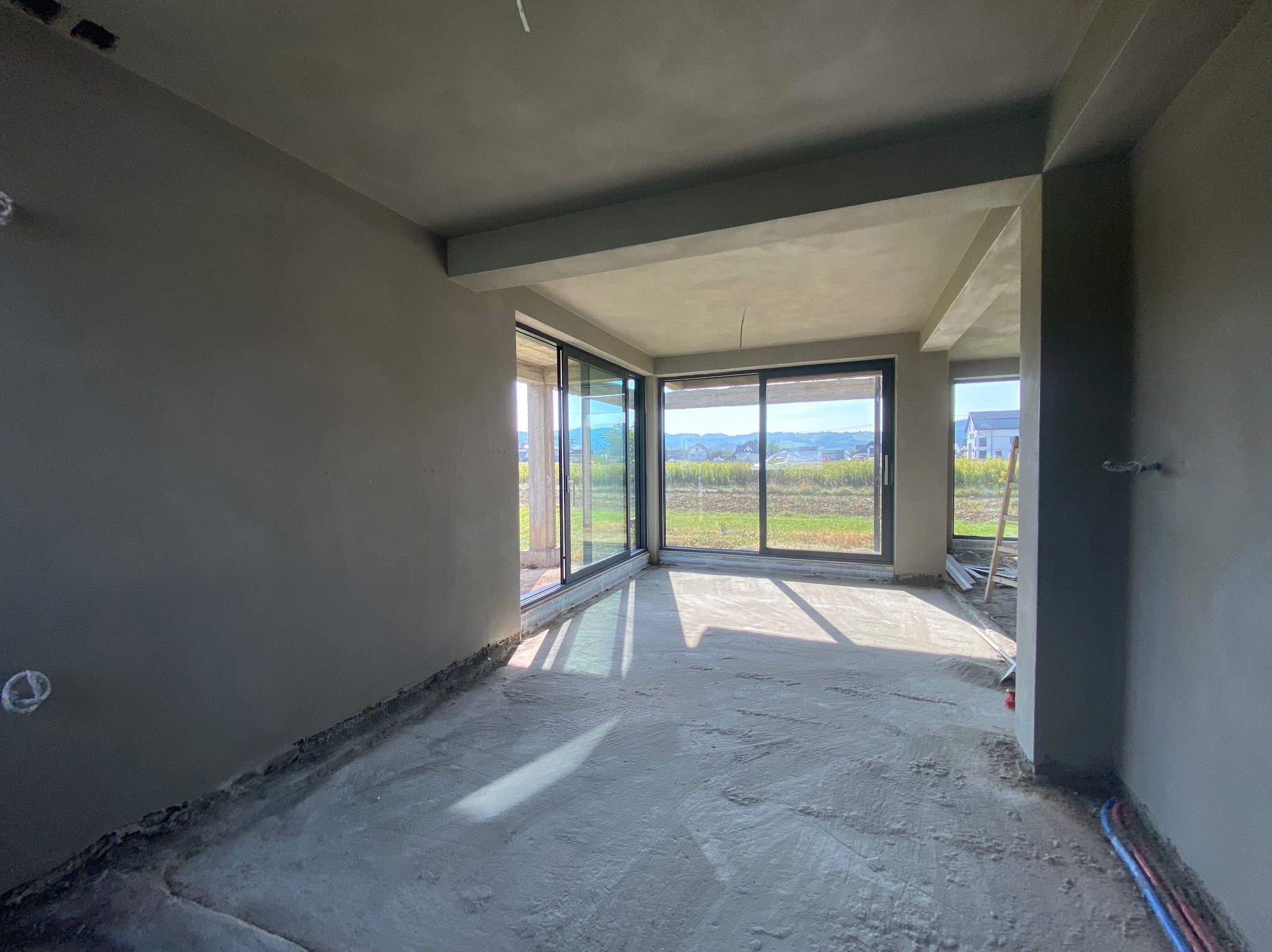 Surowe wnętrze w stanie deweloperskim z dużymi oknami przesuwnymi, ściany pokryte szarą warstwą, podłoga wylewka betonowa, widoczne instalacje przy podłodze.