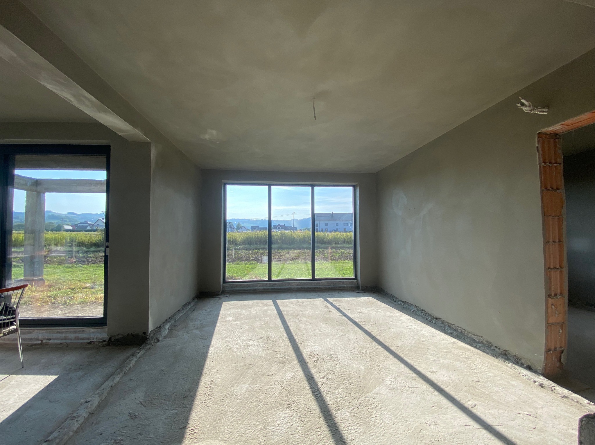 Wnętrze pokoju w stanie deweloperskim po nałożeniu gładzi, widoczne okno z widokiem na zewnątrz, surowy beton na podłodze i ściany przygotowane do dalszych prac wykończeniowych.