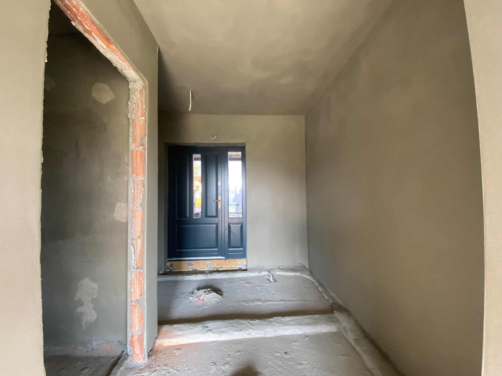 Korytarz w trakcie prac wykończeniowych: ściany i sufit pokryte świeżą warstwą szarej gładzi, widoczne surowe obramowanie drzwi po lewej stronie i ciemne drzwi wejściowe na wprost.