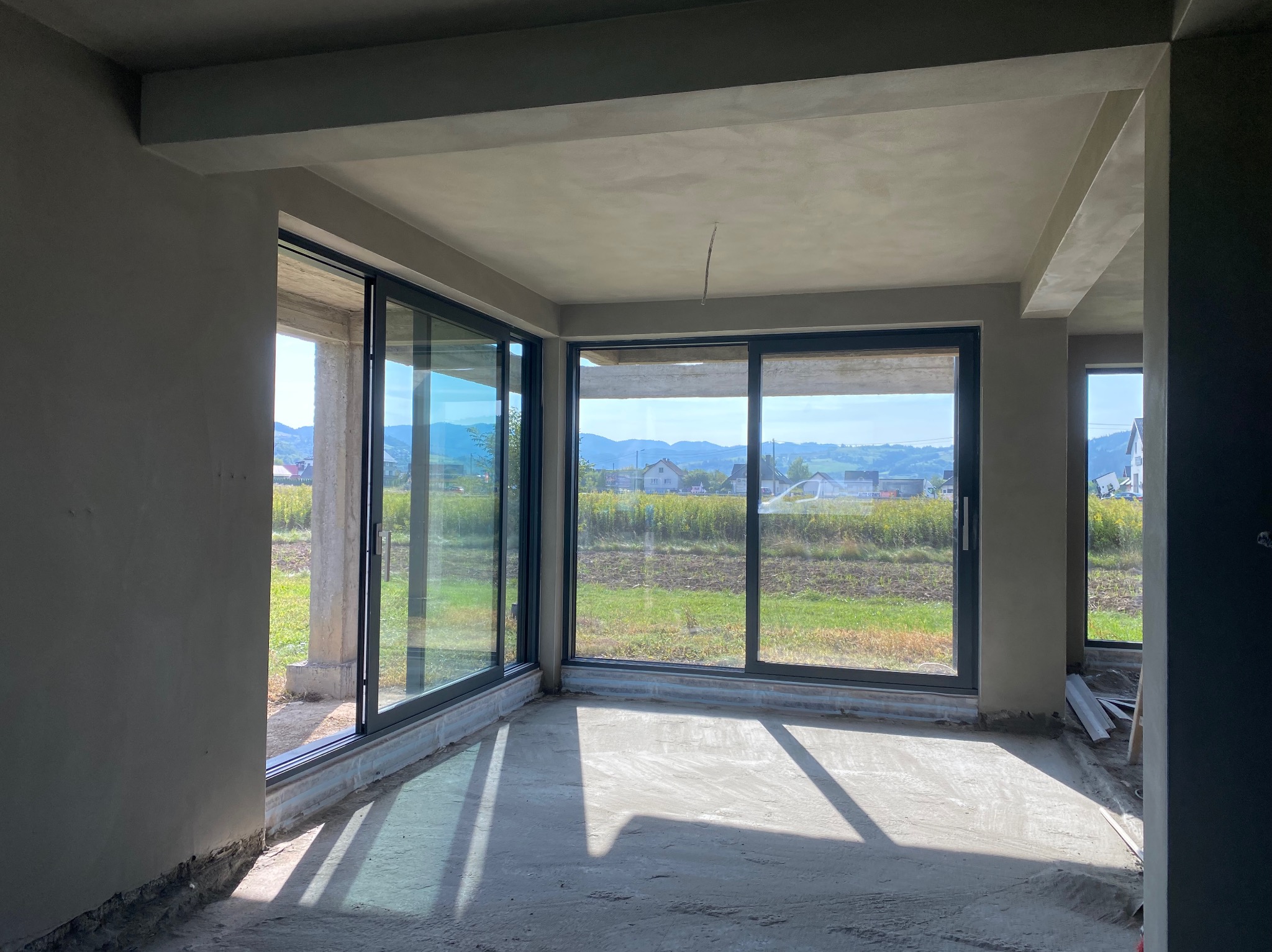 Surowe wnętrze w stanie deweloperskim z dużymi przesuwnymi oknami, szare ściany i sufit po wstępnym wykończeniu, widok na zielone pola i wzgórza za oknami.