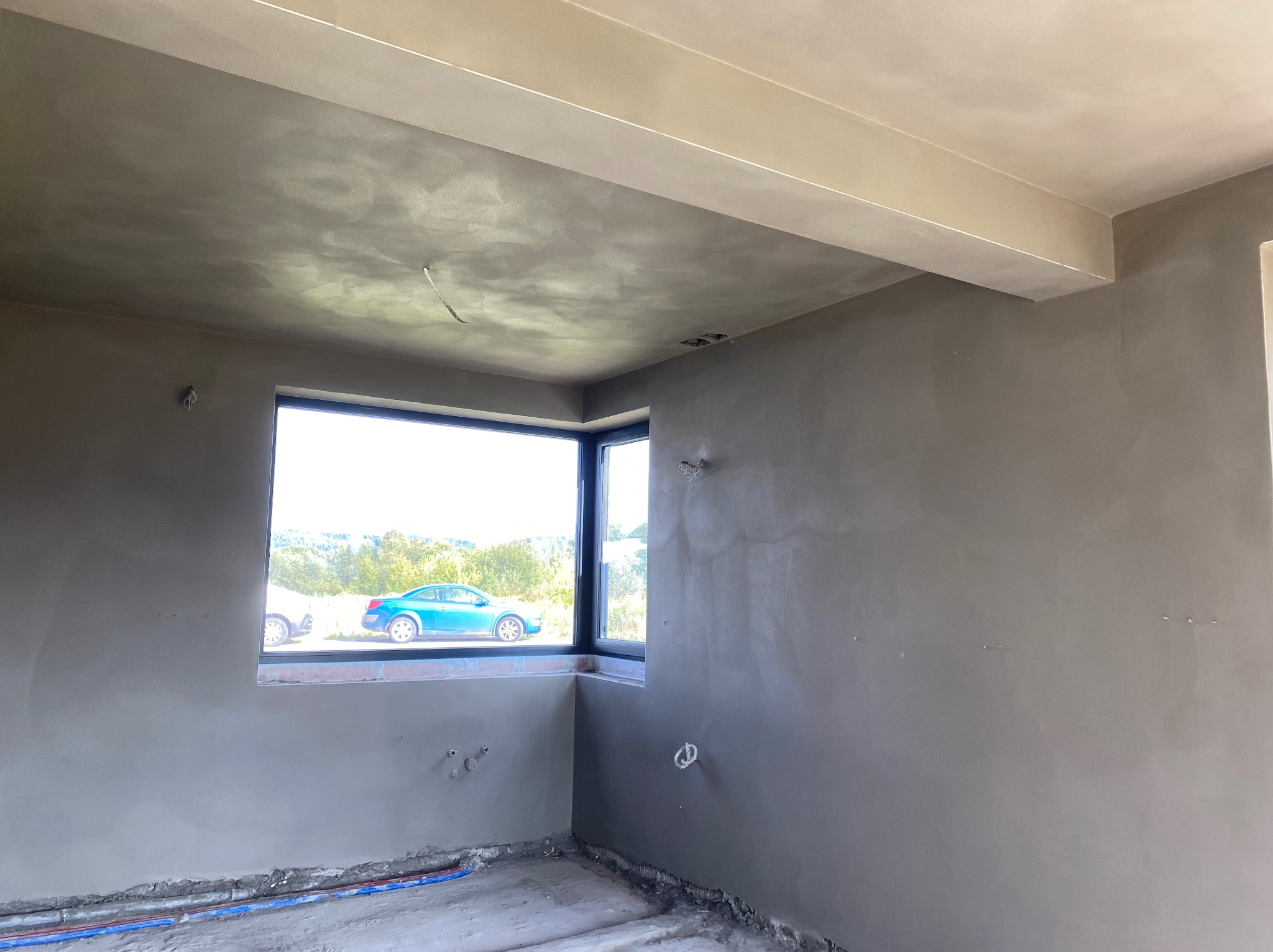 Pokój w trakcie prac wykończeniowych: świeżo nałożona szara warstwa na ścianach i suficie, widoczne wystające kable, okno z widokiem na zieleń i niebieski samochód.