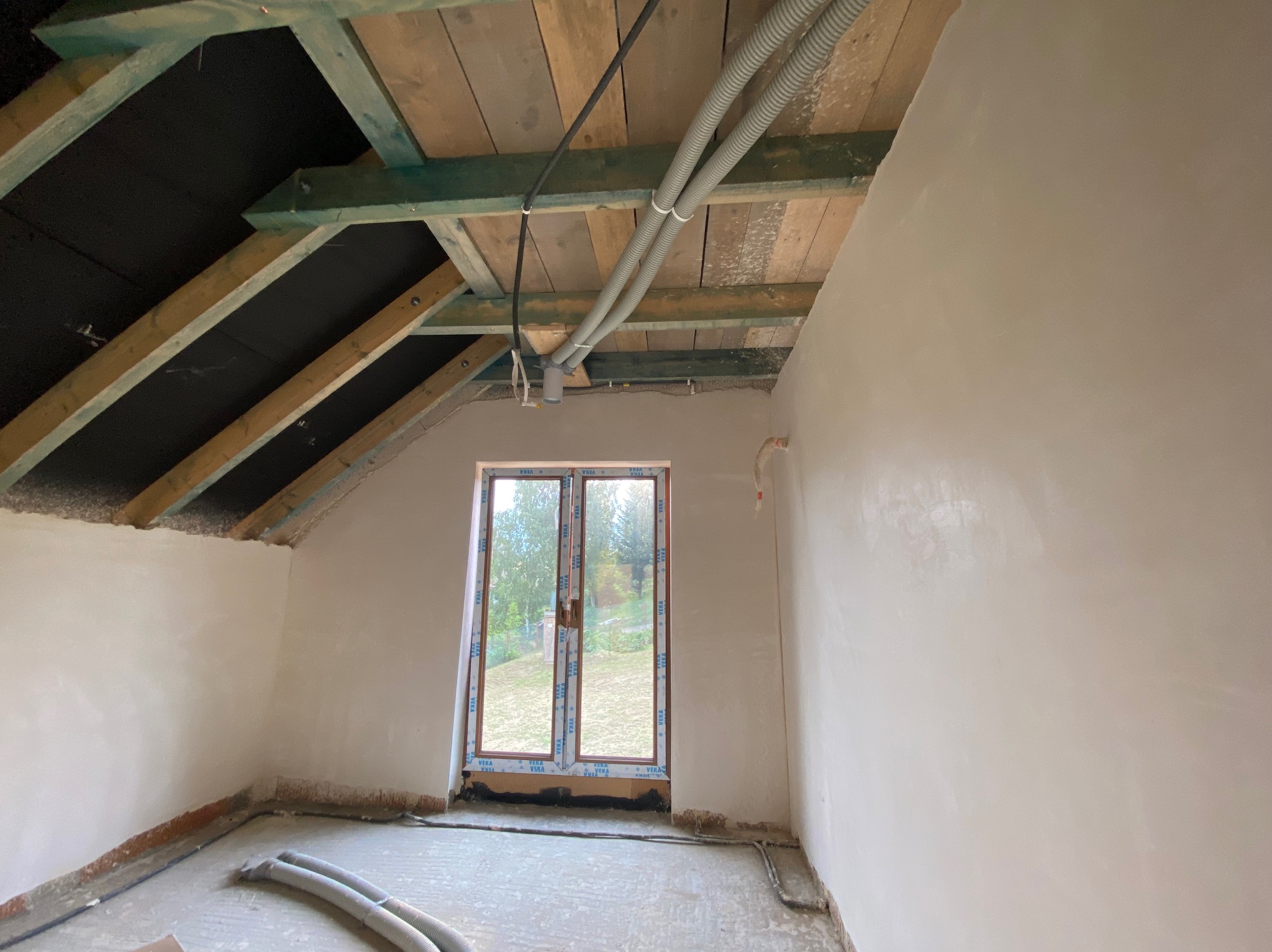 Poddasze w trakcie remontu: ściany po szpachlowaniu, widoczne rury instalacyjne pod drewnianym dachem z izolacją, okno z widokiem na zieleń.