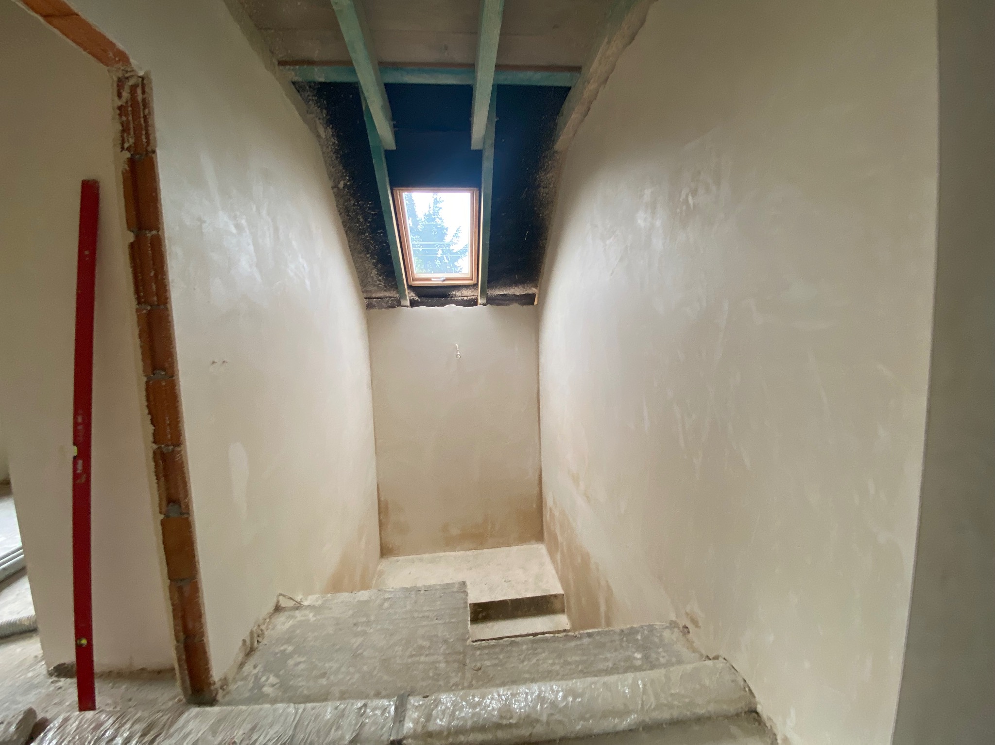 Wnętrze poddasza ze schodami w trakcie prac wykończeniowych, ściany pokryte świeżo nałożoną warstwą gładzi, widoczne okno dachowe i fragment poziomicy przy ościeżnicy.