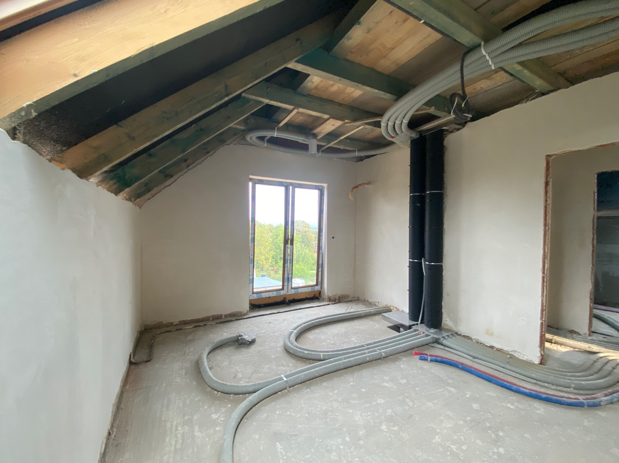 Poddasze w trakcie prac wykończeniowych: widoczne nieotynkowane ściany, drewniana konstrukcja dachu z poprowadzonymi przewodami wentylacyjnymi i elektrycznymi, okno z widokiem na zieleń.