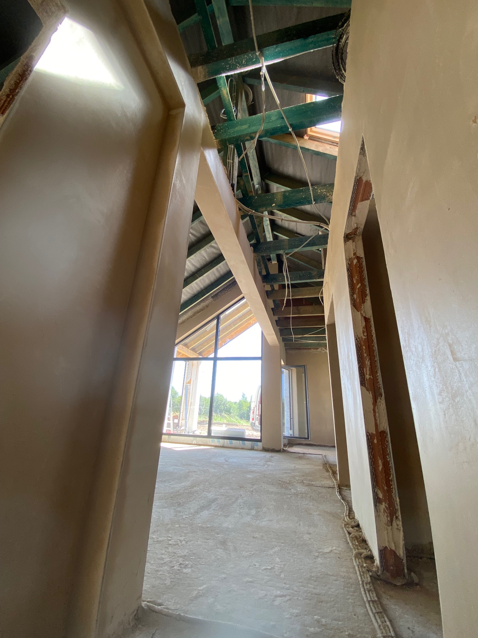 Wnętrze w trakcie prac wykończeniowych: surowe ściany po nałożeniu warstwy wyrównującej, widoczna drewniana konstrukcja dachu z oknem dachowym, kable elektryczne prowadzone w podłodze.