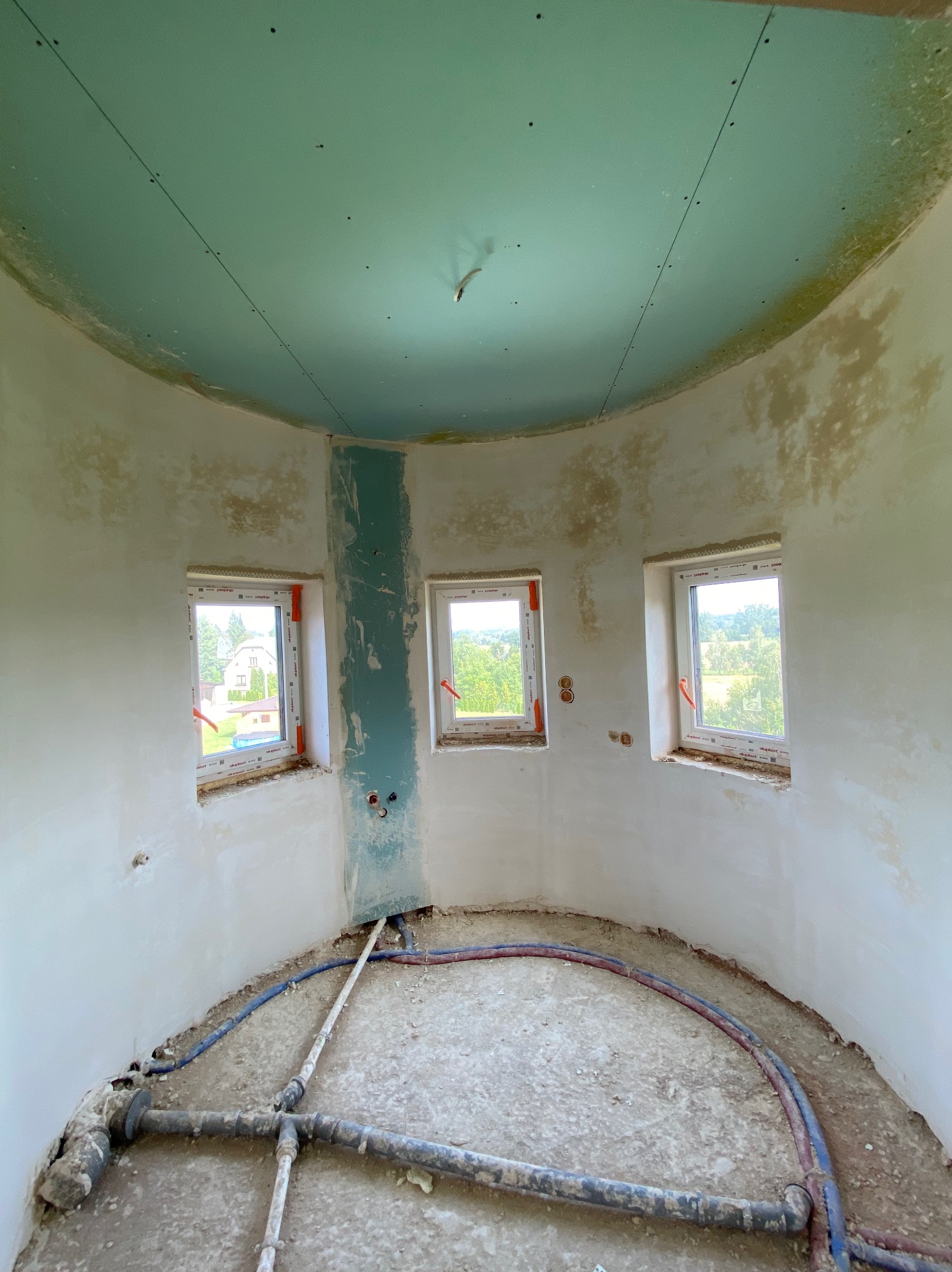 Okrągłe pomieszczenie w trakcie prac wykończeniowych: sufit pokryty zielonkawą płytą, ściany częściowo wyszpachlowane, trzy okna z widokiem na zieleń, rury na podłodze.