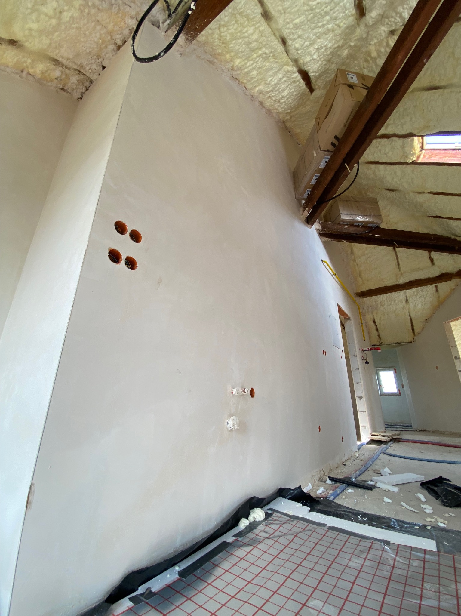 Ściana poddasza po nałożeniu gładzi, z widocznymi otworami na gniazdka elektryczne i instalacją ogrzewania podłogowego w trakcie montażu, drewniane belki stropowe z pianką izolacyjną i kartonami...