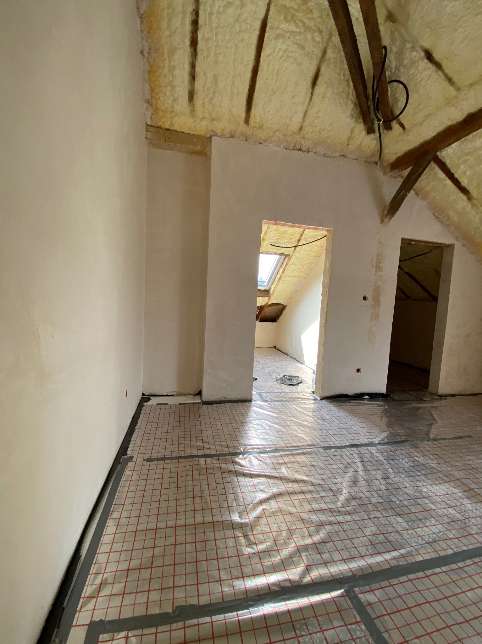 Poddasze w trakcie prac wykończeniowych: ściany po położeniu gładzi, izolacja natryskowa dachu widoczna, na podłodze folia grzewcza z czerwoną siatką.