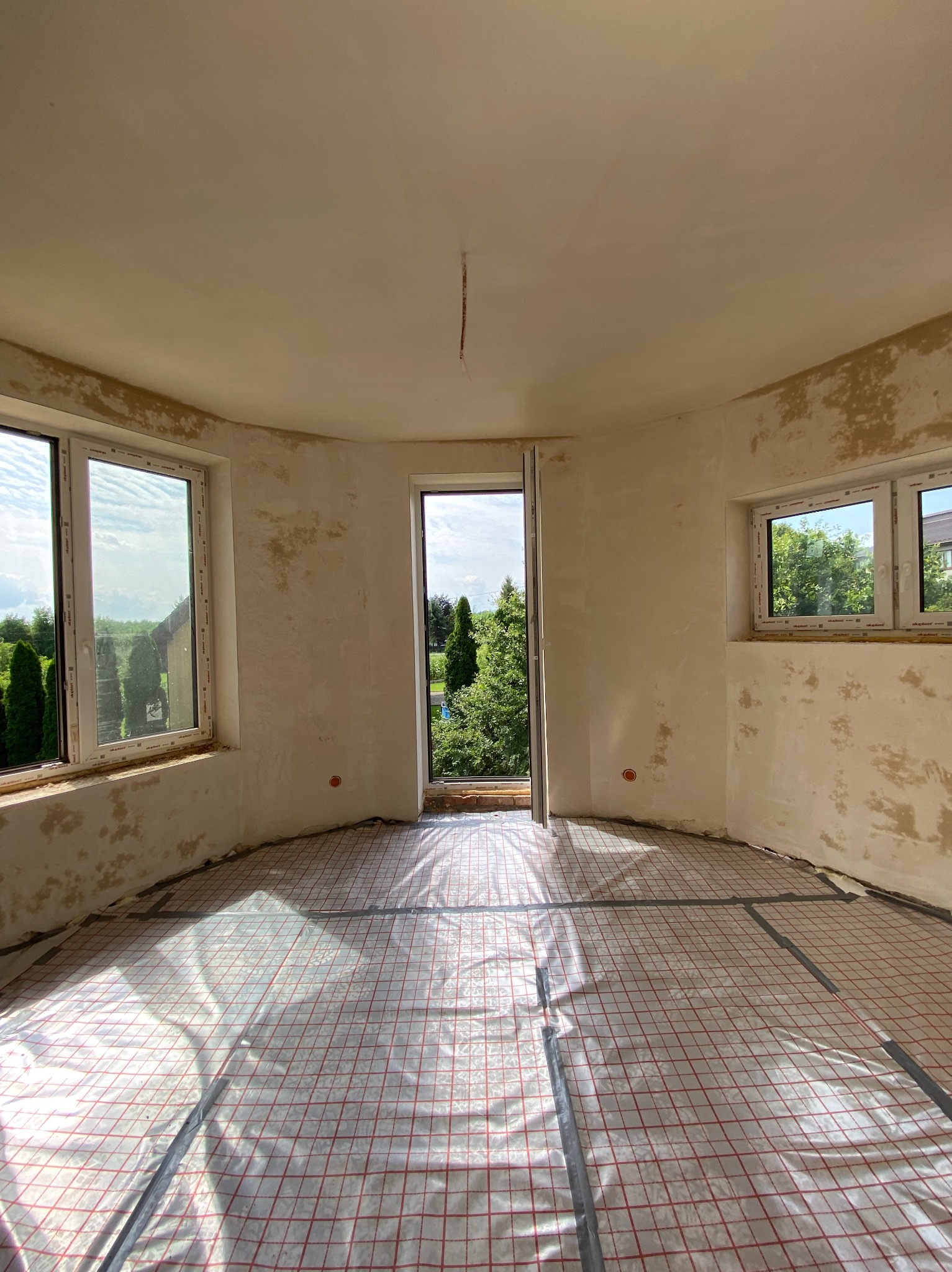 Wnętrze okrągłego pokoju w trakcie prac przygotowawczych: ściany z surową powierzchnią, podłoga zabezpieczona folią z czerwoną kratką, widok na zieleń przez okna i otwarte drzwi.