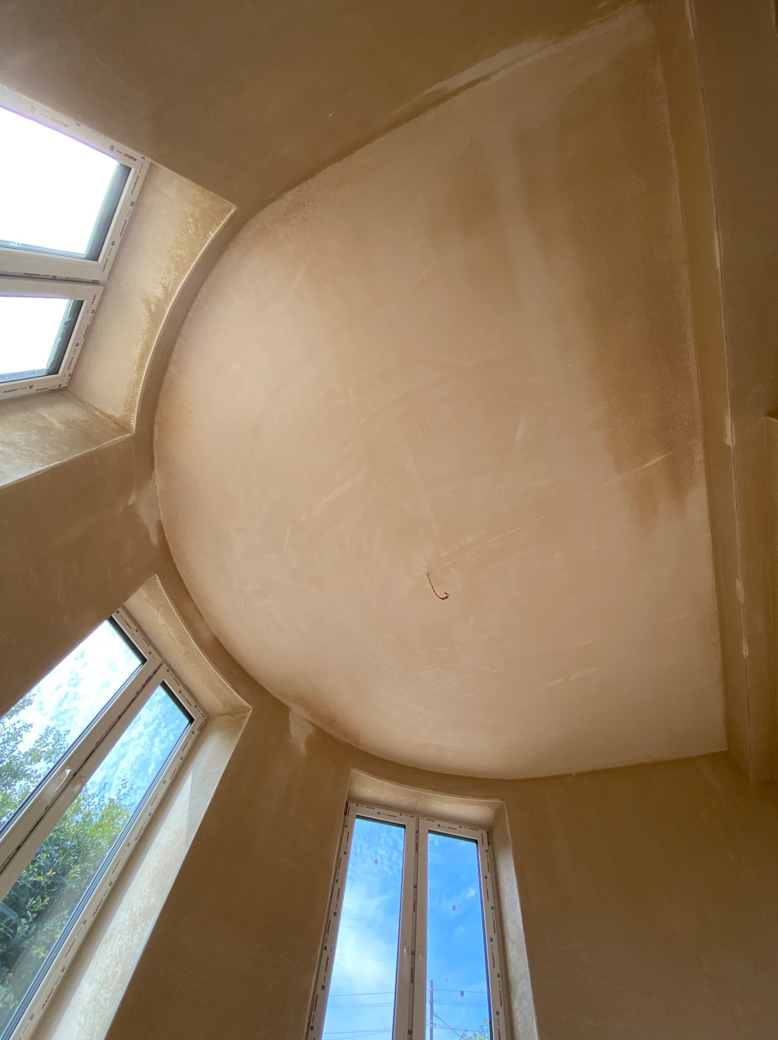 Okrągłe sklepienie sufitowe w trakcie prac wykończeniowych, widoczne świeżo nałożone warstwy, trzy okna z widokiem na zewnątrz, ślady niedoskonałości na powierzchni.