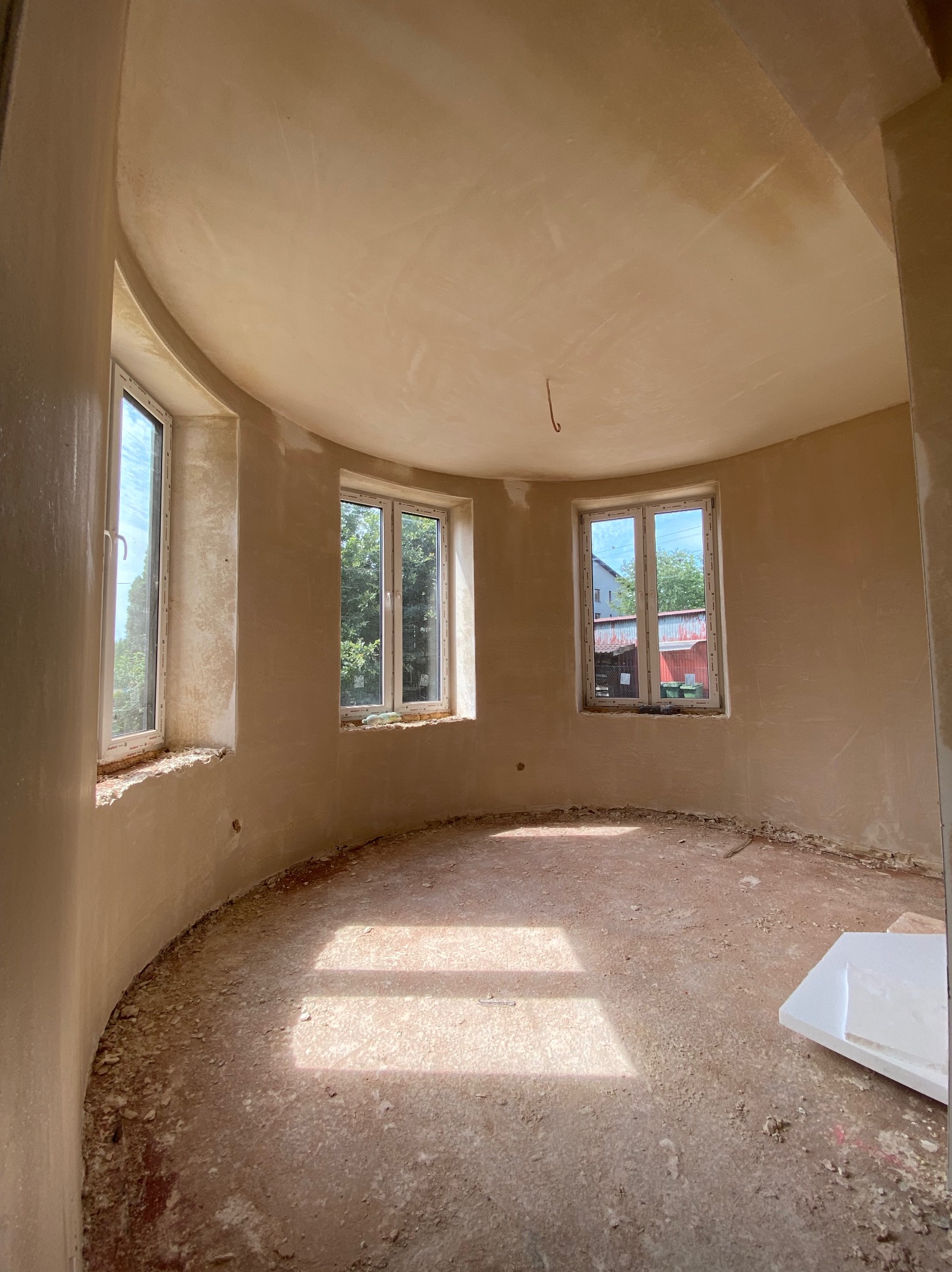 Okrągłe pomieszczenie w trakcie prac wykończeniowych; ściany pokryte świeżo nałożoną warstwą wygładzającą, widoczne trzy okna z widokiem na zieleń, podłoga z resztkami materiałów budowlanych.