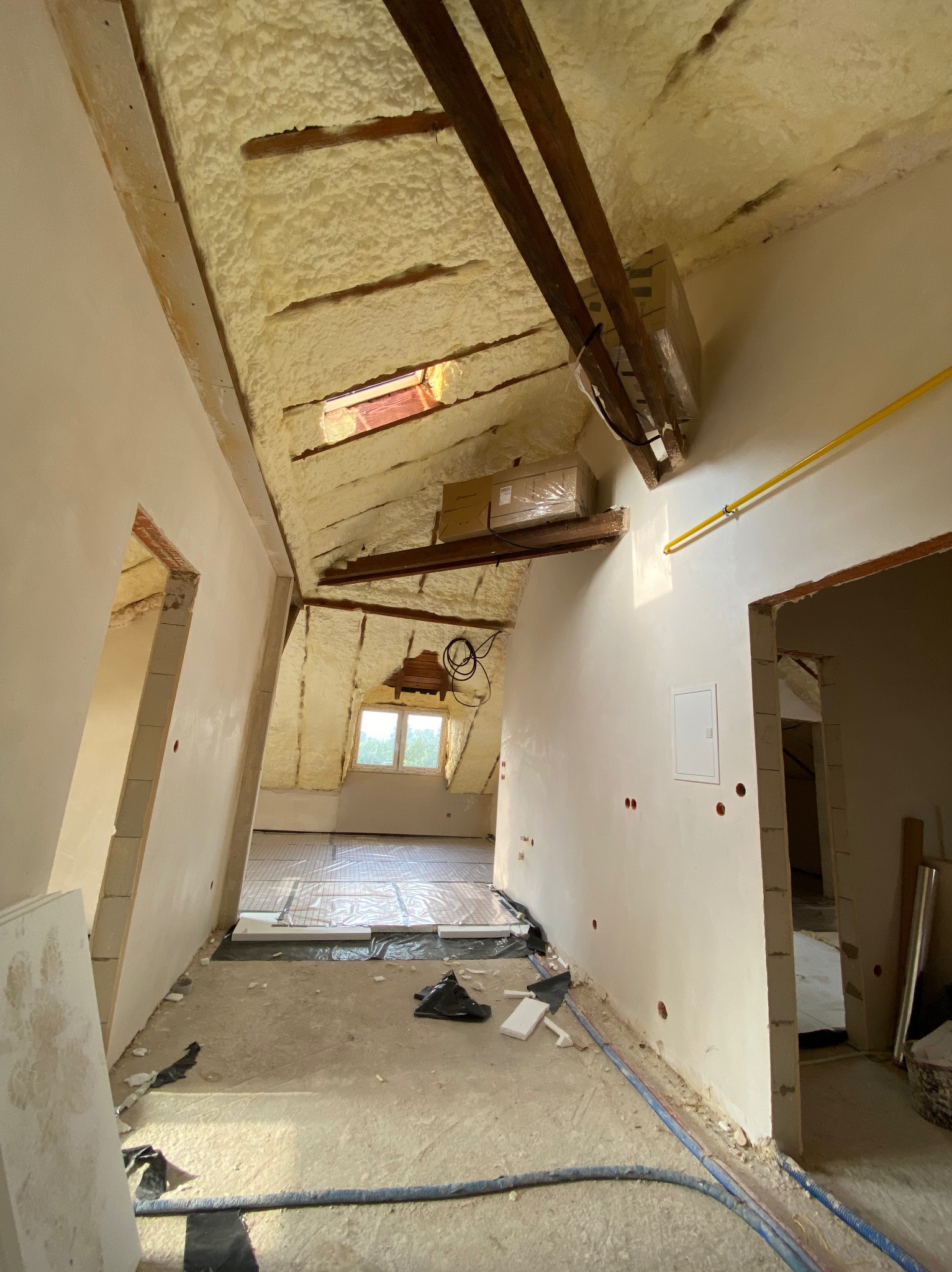 Poddasze w trakcie remontu: izolacja pianą PUR na skośnym dachu z widocznymi belkami, instalacja ogrzewania podłogowego, surowe ściany po szpachlowaniu z widocznymi punktami elektrycznymi...
