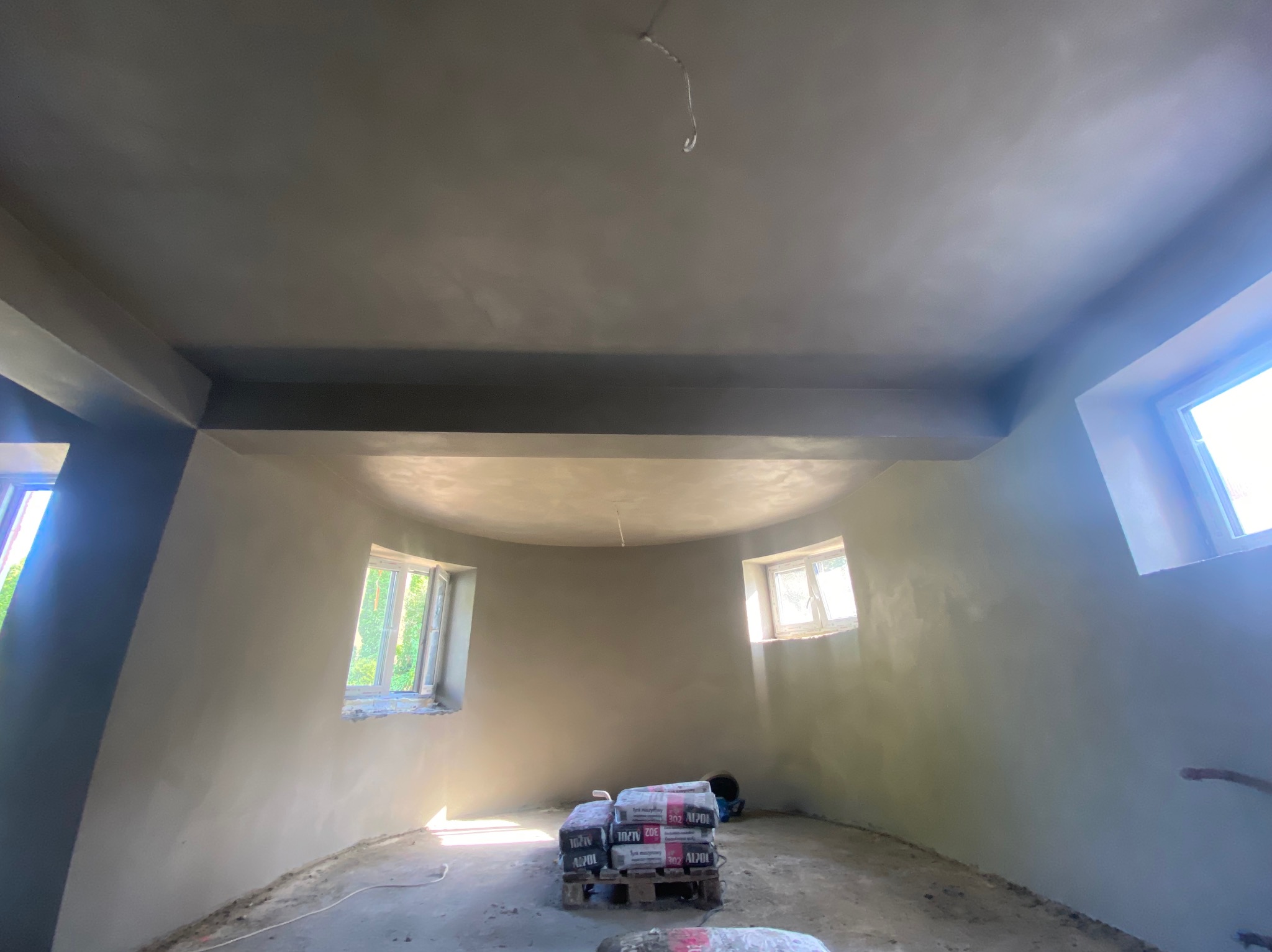Wnętrze pokoju po szpachlowaniu ścian i sufitu, widoczne worki z zaprawą na palecie, trzy okna wpuszczające naturalne światło, przewód zwisający z sufitu.