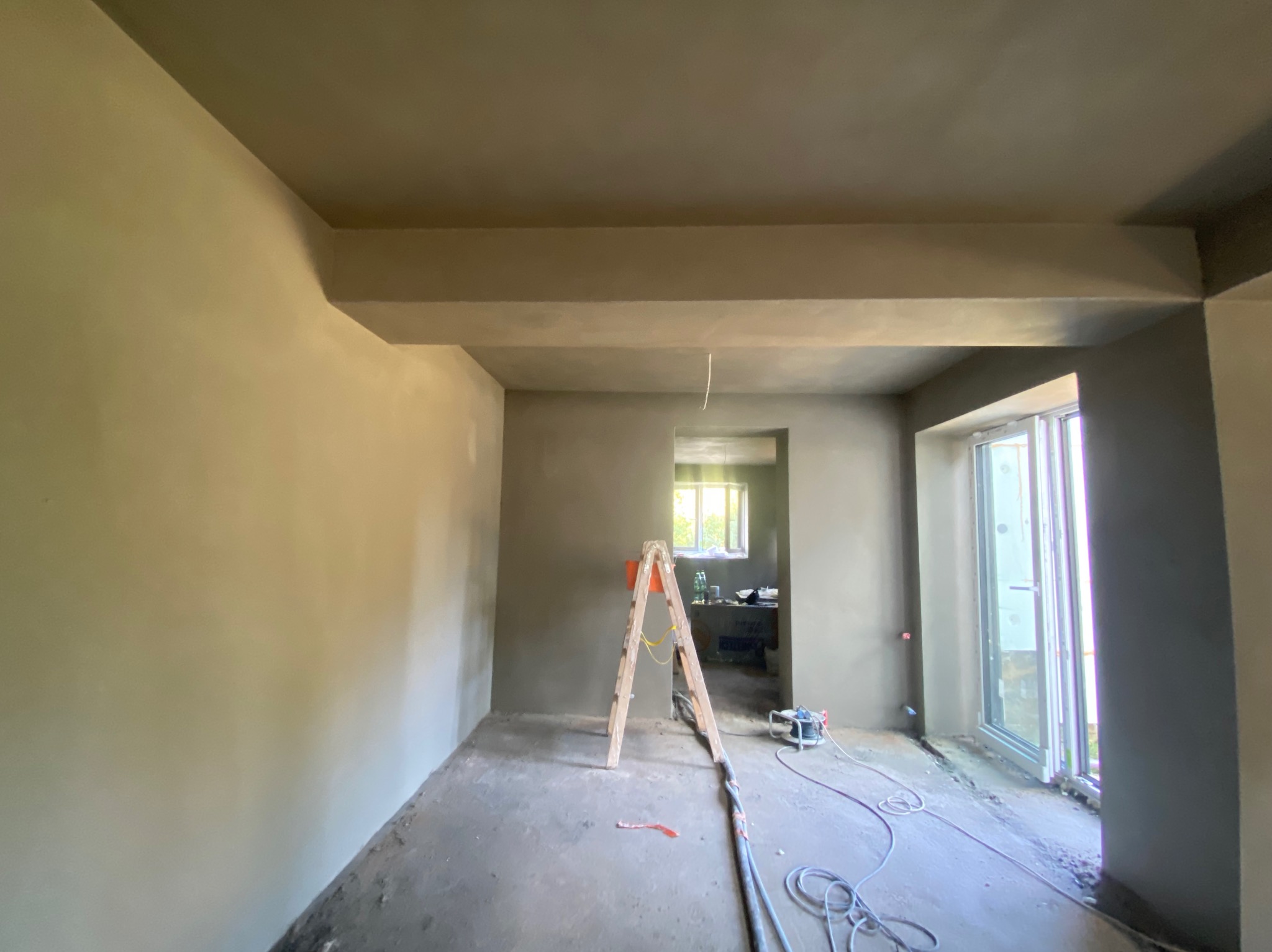 Pokój w trakcie prac wykończeniowych, ściany i sufit pokryte świeżą warstwą szpachli, widoczna drabina i przewody urządzenia do natrysku, w tle okno z widokiem na zieleń.