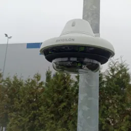 Kamera przemysłowa Avigilon zamontowana na metalowym słupie, na tle zielonych drzew i hali magazynowej w pochmurny dzień.