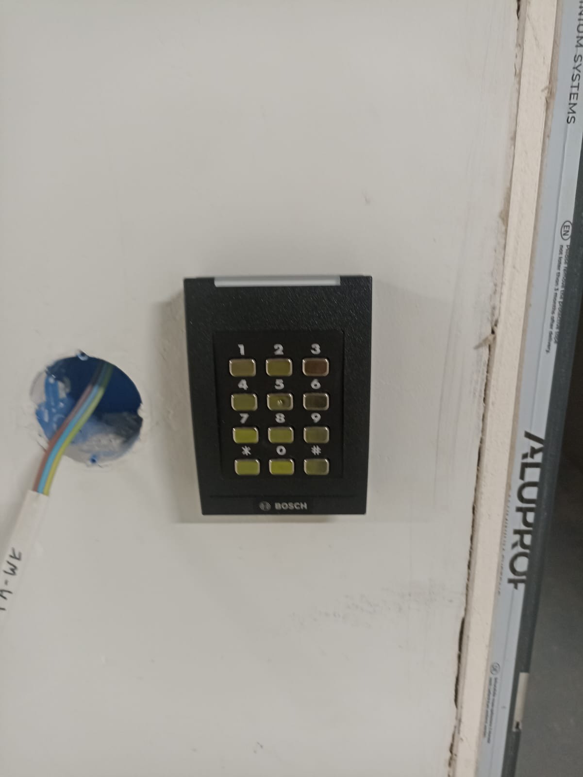Klawiatura numeryczna Bosch zamontowana na ścianie obok otworu z wystającymi przewodami, widoczny fragment ramy okiennej.