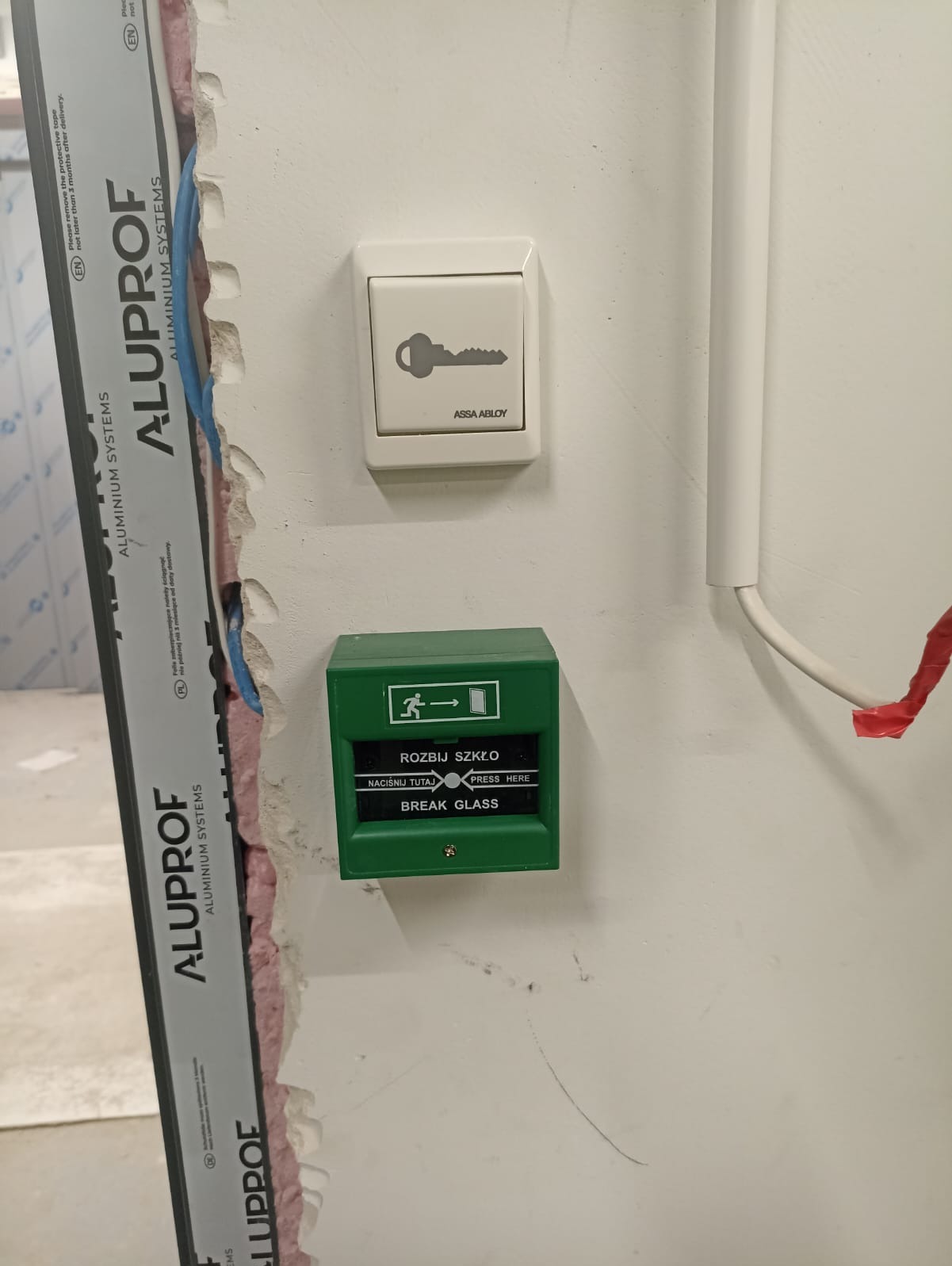 Biały wyłącznik z symbolem klucza i zielony przycisk alarmowy 'Rozbij szkło' zamontowane na ścianie, widoczne kable w ścianie.