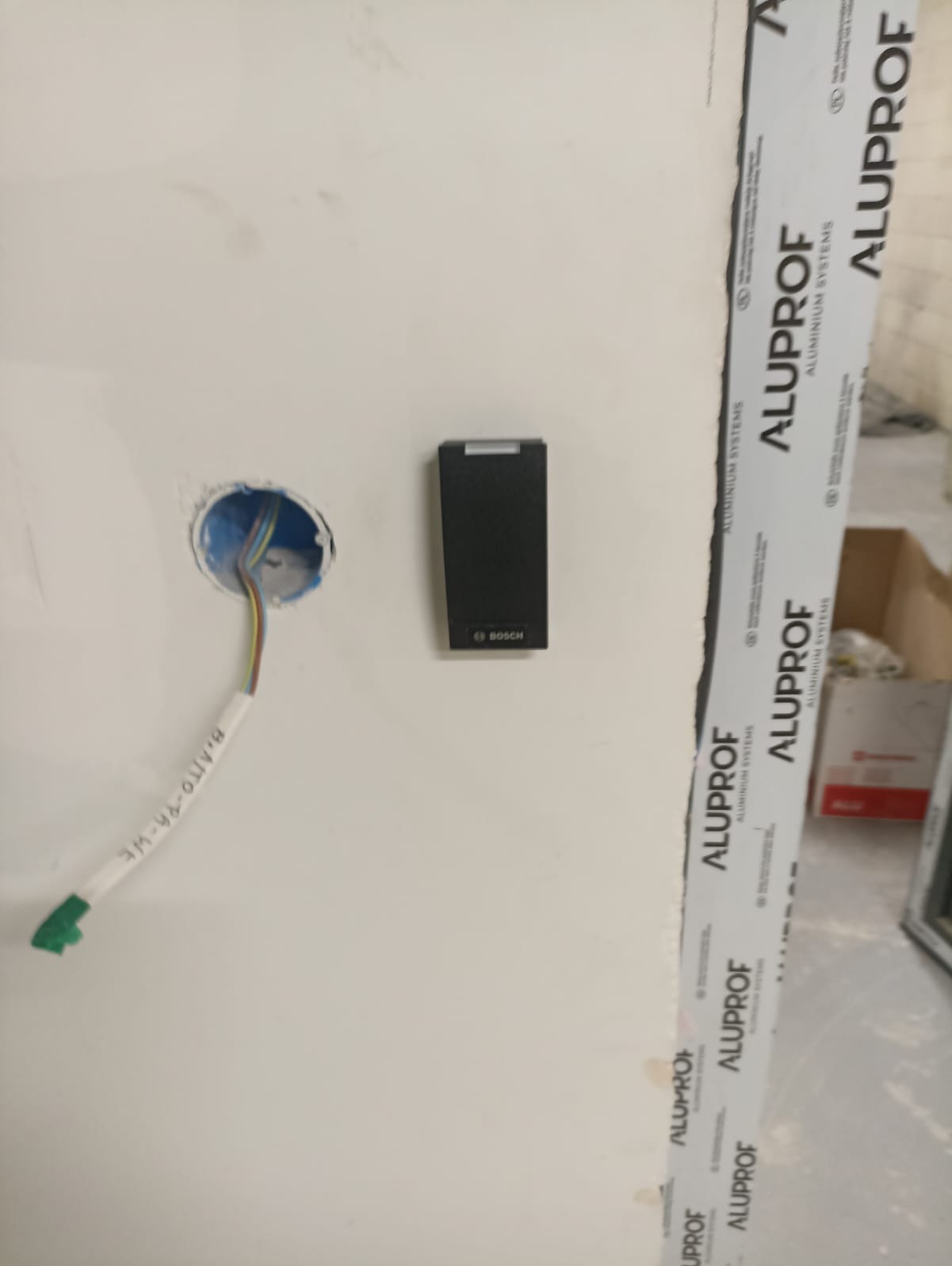 Montaż czujnika alarmowego Bosch na ścianie obok otworu z przewodami elektrycznymi, widoczny profil okienny Aluprof.