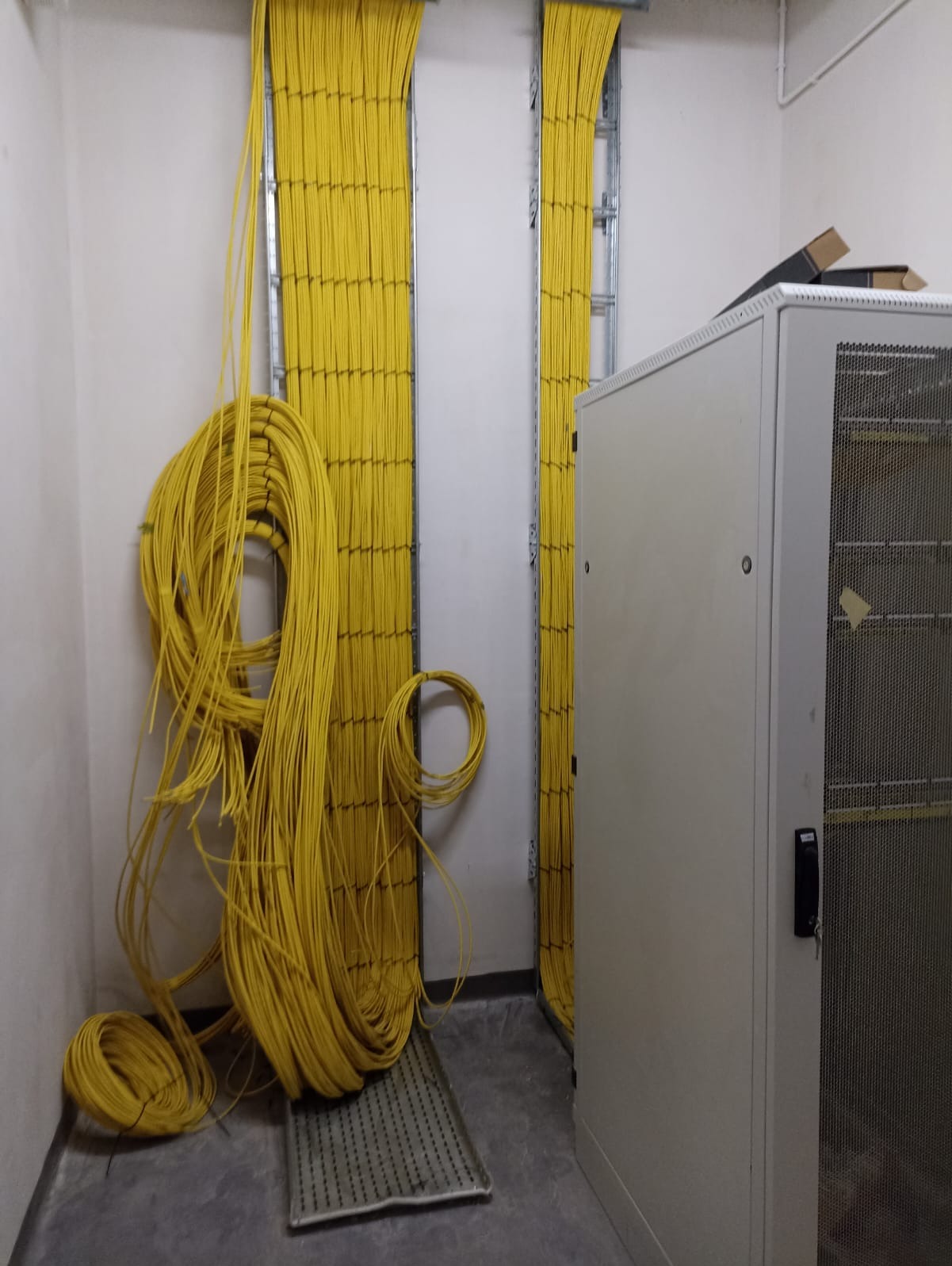 Żółte kable sieciowe ułożone w korytach kablowych w pomieszczeniu technicznym, część kabli zwisa luźno na podłodze obok kratki wentylacyjnej, obok szafa rack.
