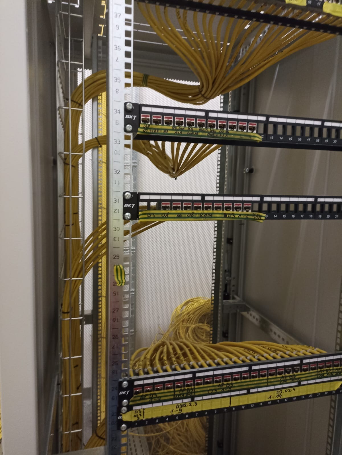 Panel krosowniczy z żółtymi kablami sieciowymi w szafie rack, widoczne oznaczenia na kablach i panelach, metalowa konstrukcja szafy.