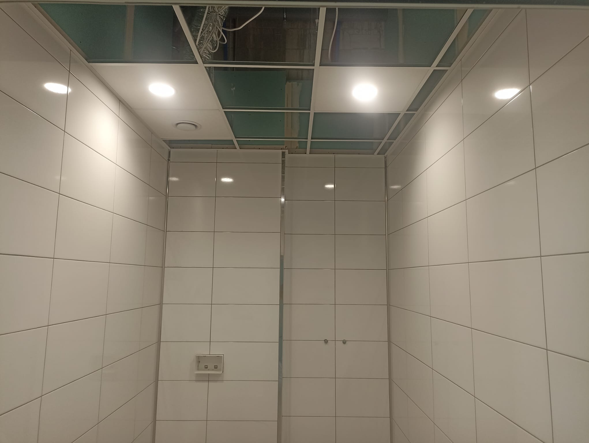 Łazienka w trakcie remontu: białe kafelki na ścianach, sufit podwieszany z widoczną instalacją elektryczną i punktowymi lampami, brakujące panele sufitowe.