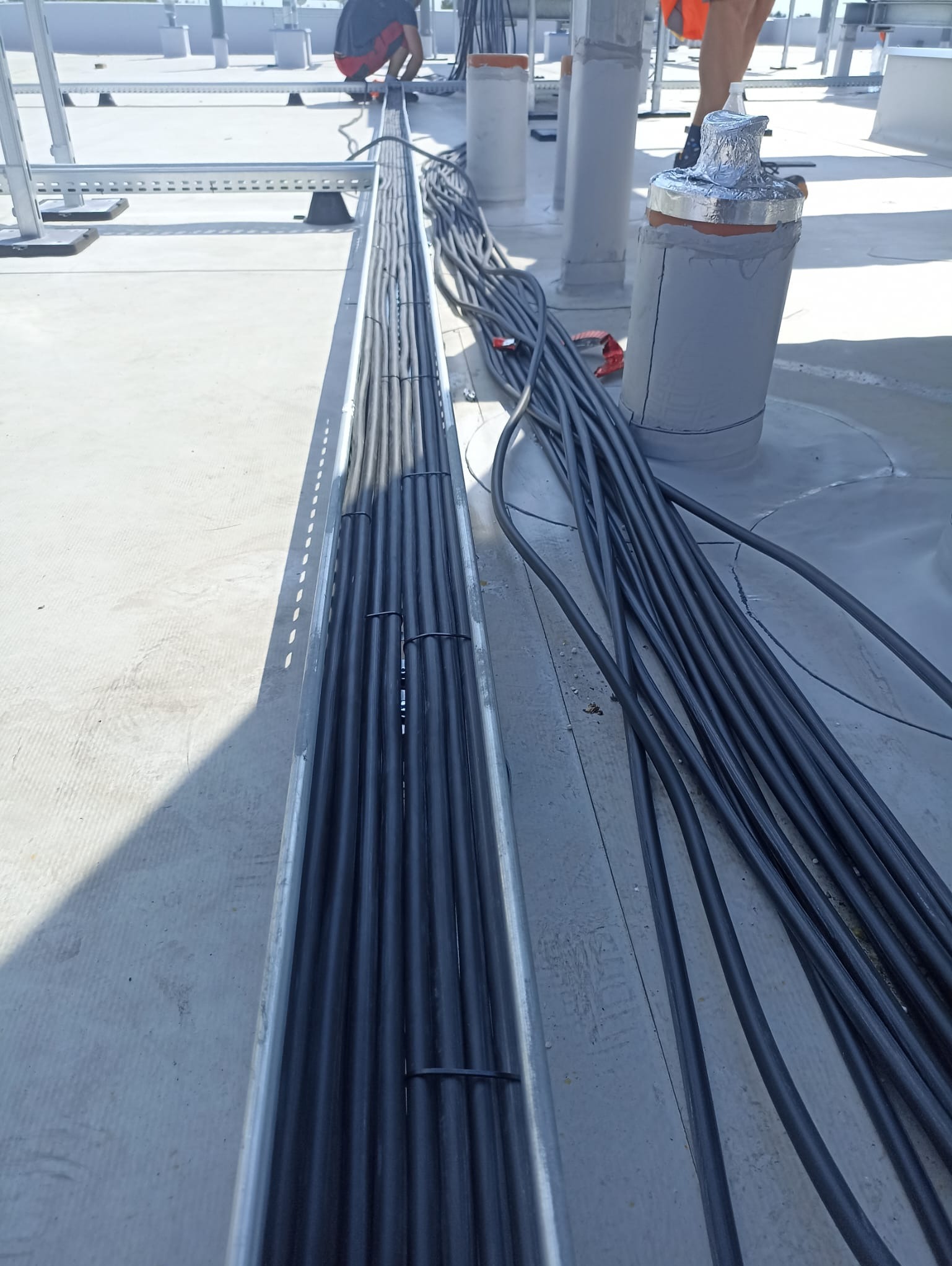 Instalacja kabli elektrycznych w korycie na dachu, widoczne zabezpieczone przepusty instalacyjne i pracownicy w tle.