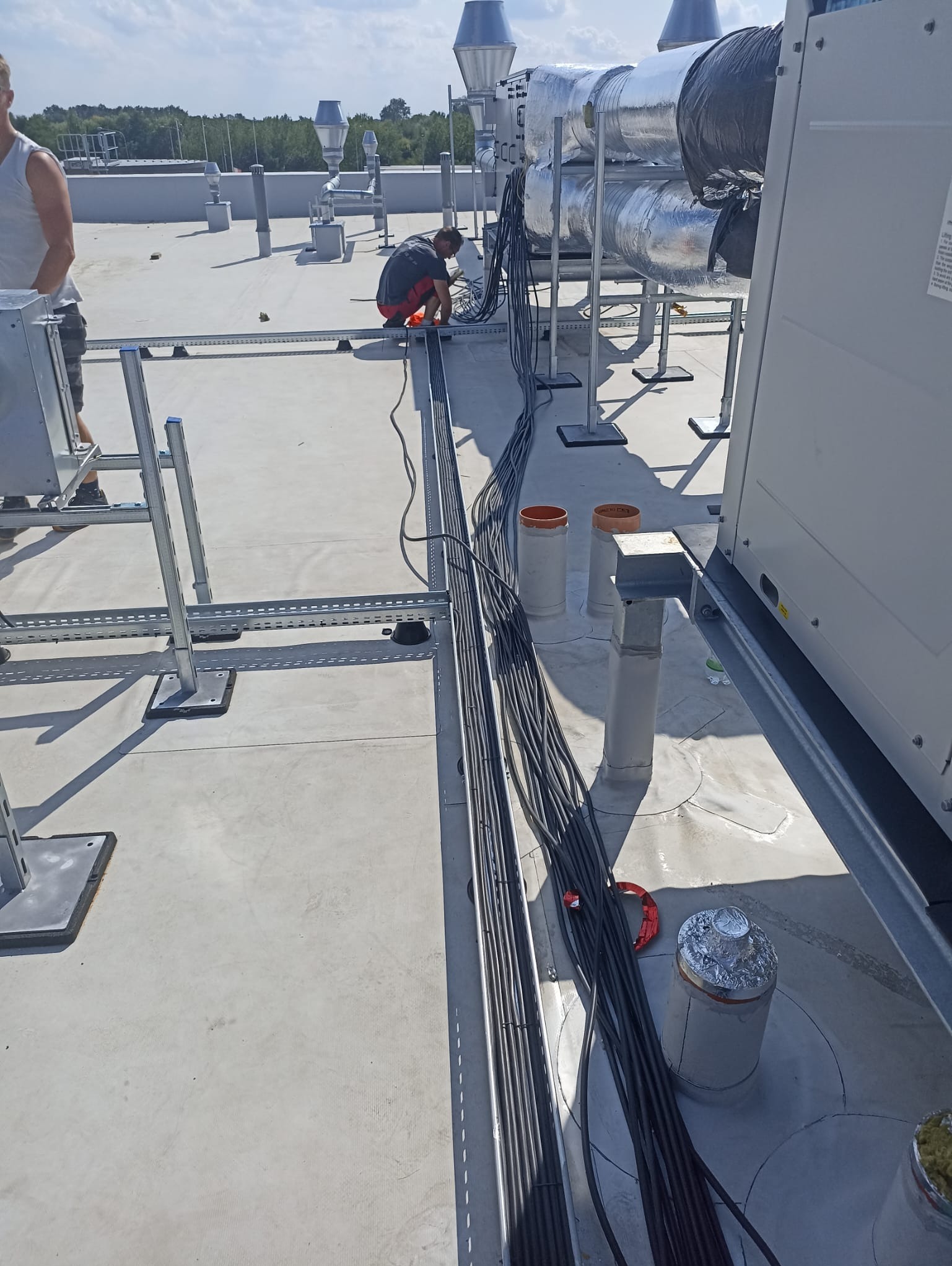 Instalacja przewodów elektrycznych na dachu budynku przemysłowego, widoczny pracownik w trakcie pracy, koryta kablowe i system wentylacji.