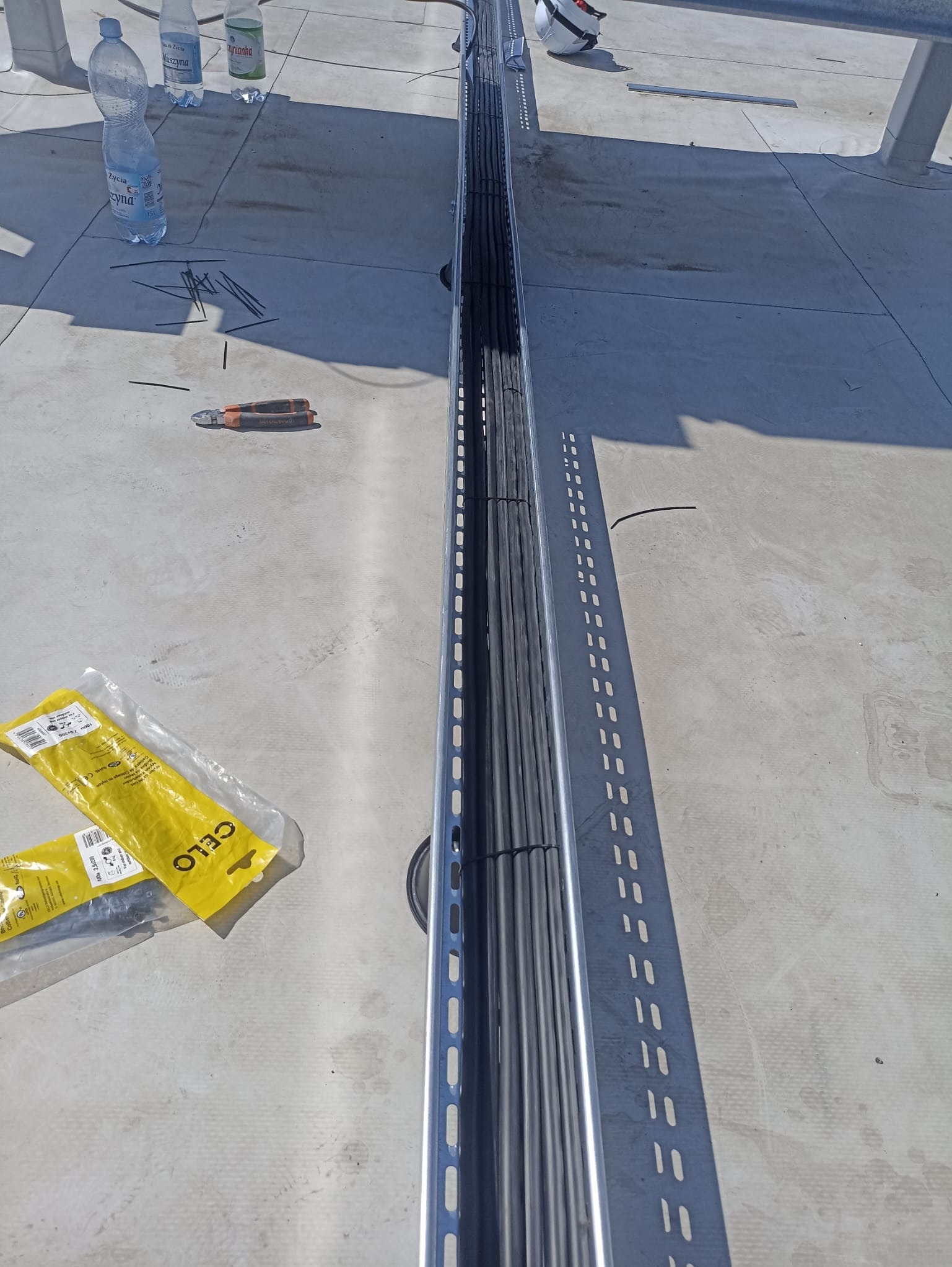 Metalowa korytka kablowa wypełniona czarnymi kablami na szarym dachu, obok leżą butelki wody, obcinaczki do kabli i opakowania opasek zaciskowych CELO.