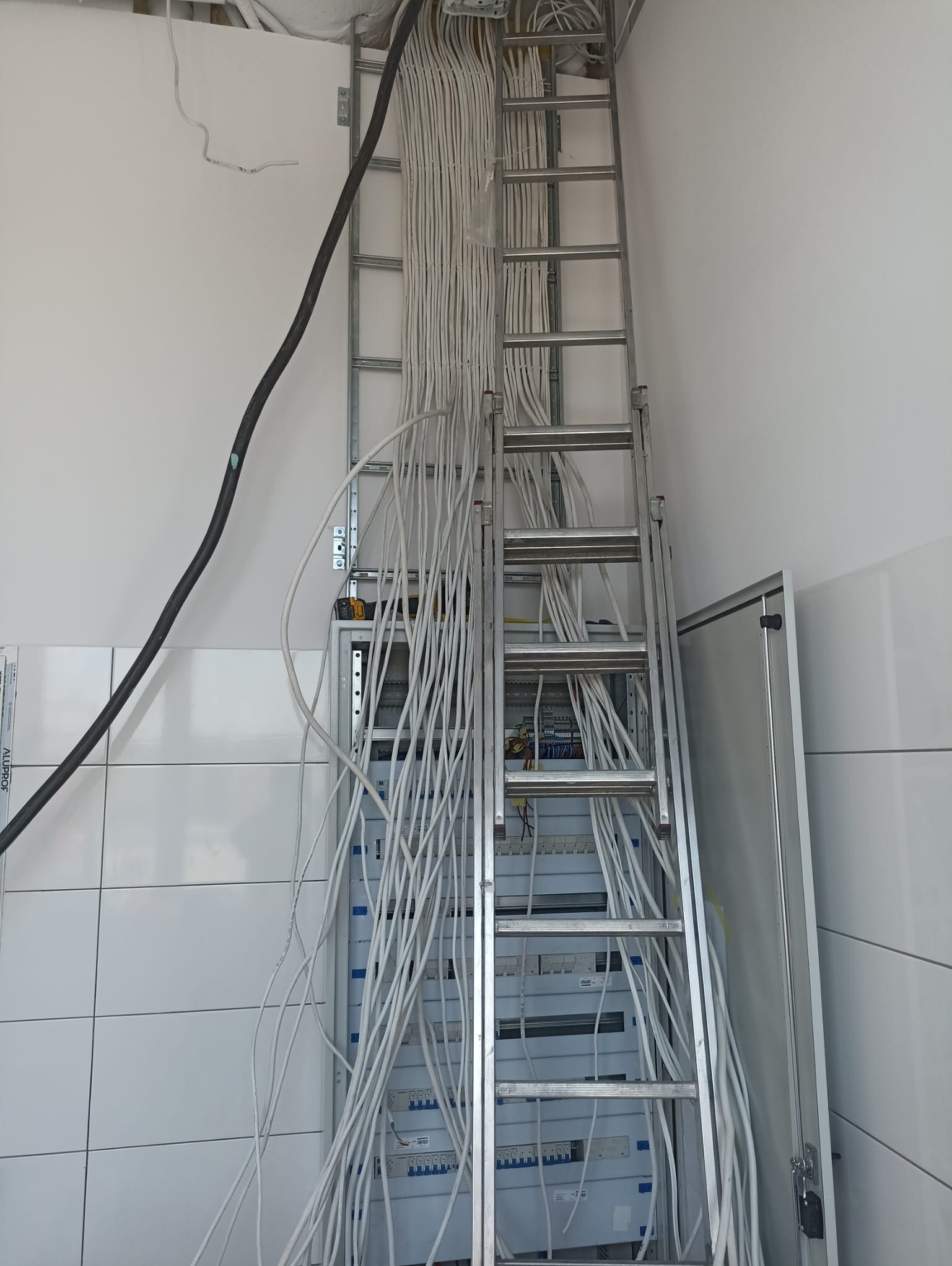 Rozdzielnica elektryczna z licznymi przewodami instalacyjnymi, drabina aluminiowa oparta o ścianę, widoczne narzędzia i fragment czarnego kabla zasilającego.