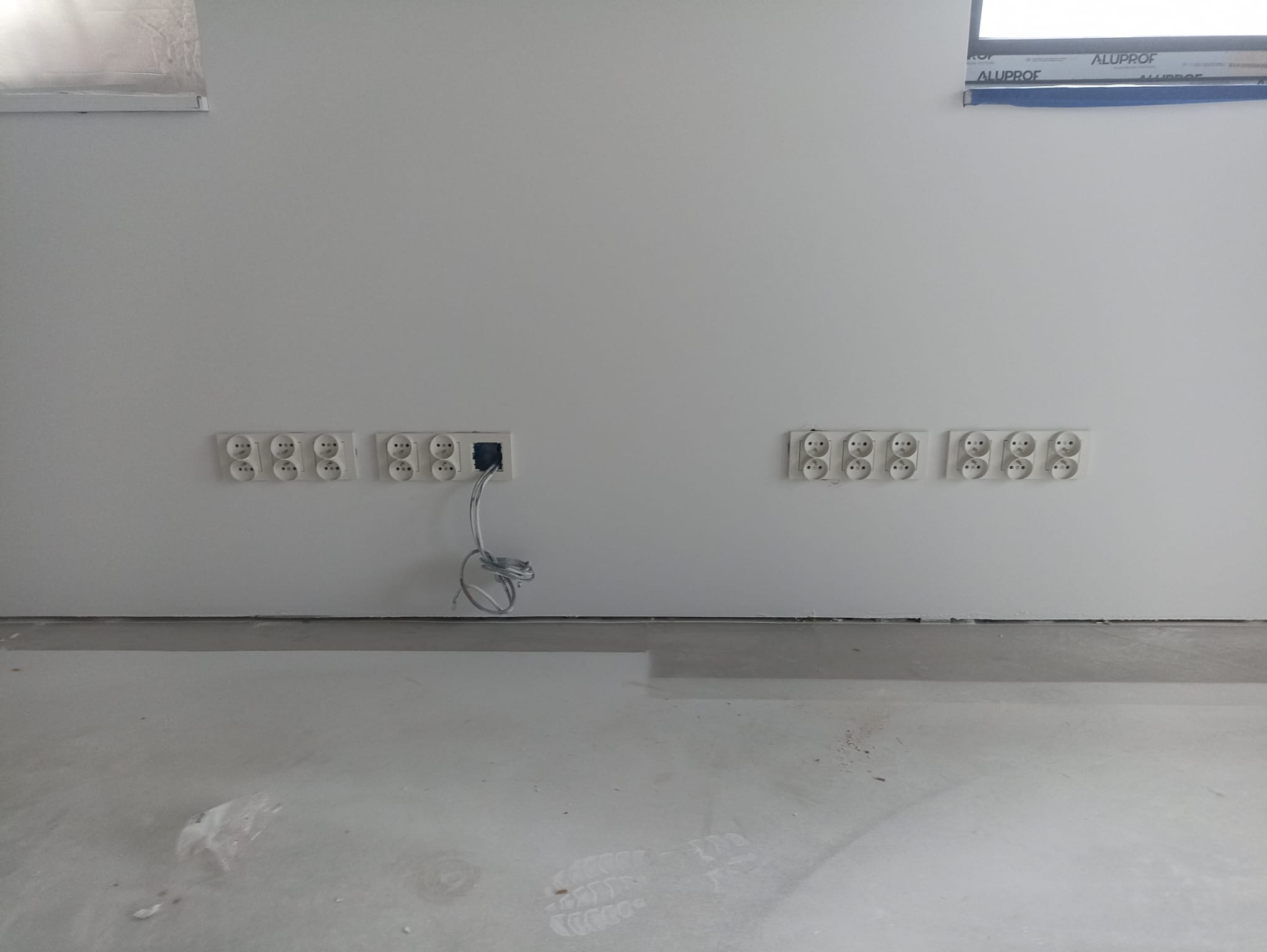 Równomiernie rozmieszczone gniazdka elektryczne w rzędach na świeżo pomalowanej ścianie, przewody wystające z jednego gniazda, surowy beton na podłodze, widoczne ślady po pracach wykończeniowych.