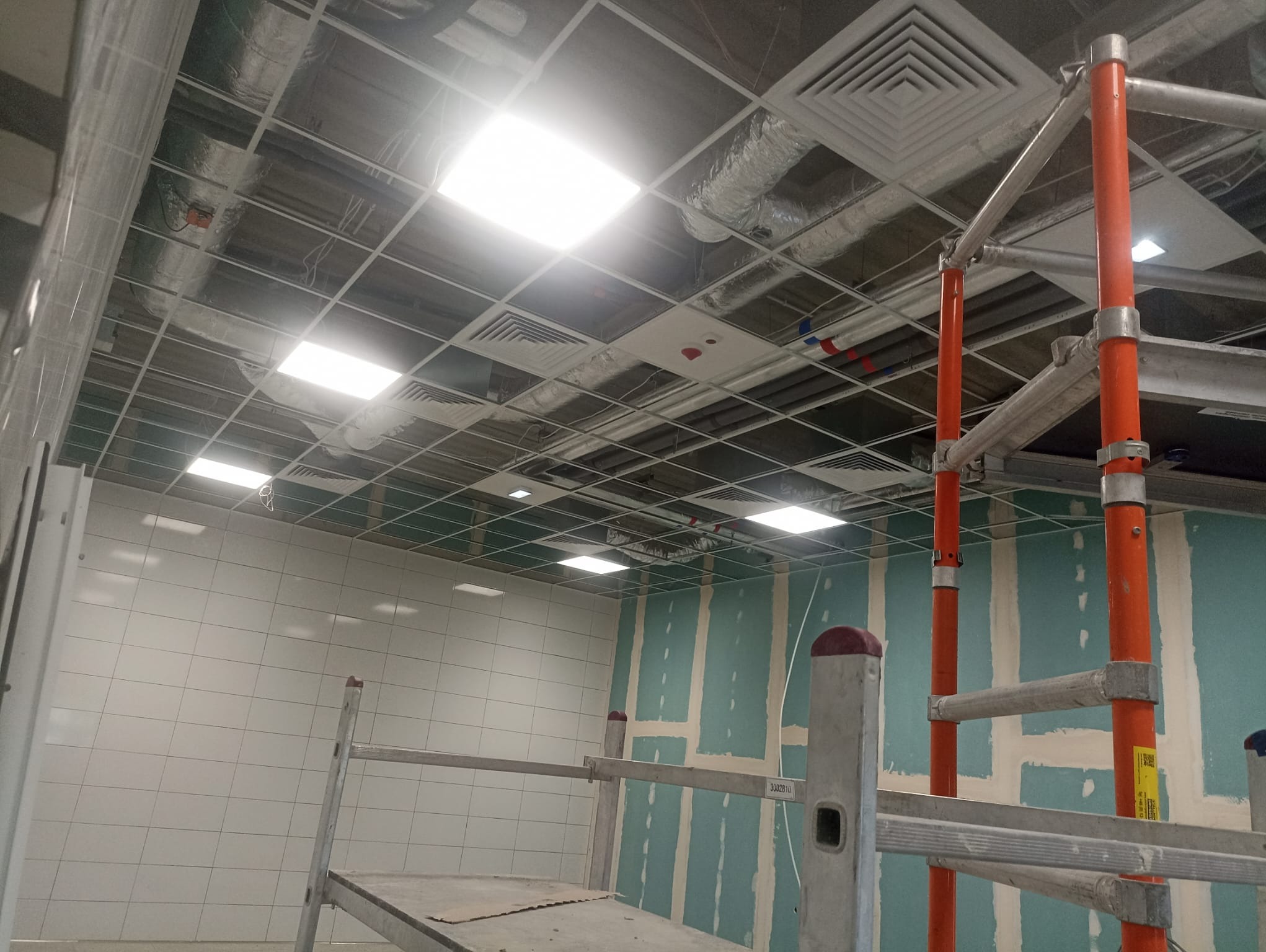 Wnętrze w trakcie prac wykończeniowych: widoczne rury wentylacyjne pod sufitem, rusztowanie, ściany z płyt gipsowo-kartonowych przygotowane do malowania i częściowo wyłożone białymi płytkami.