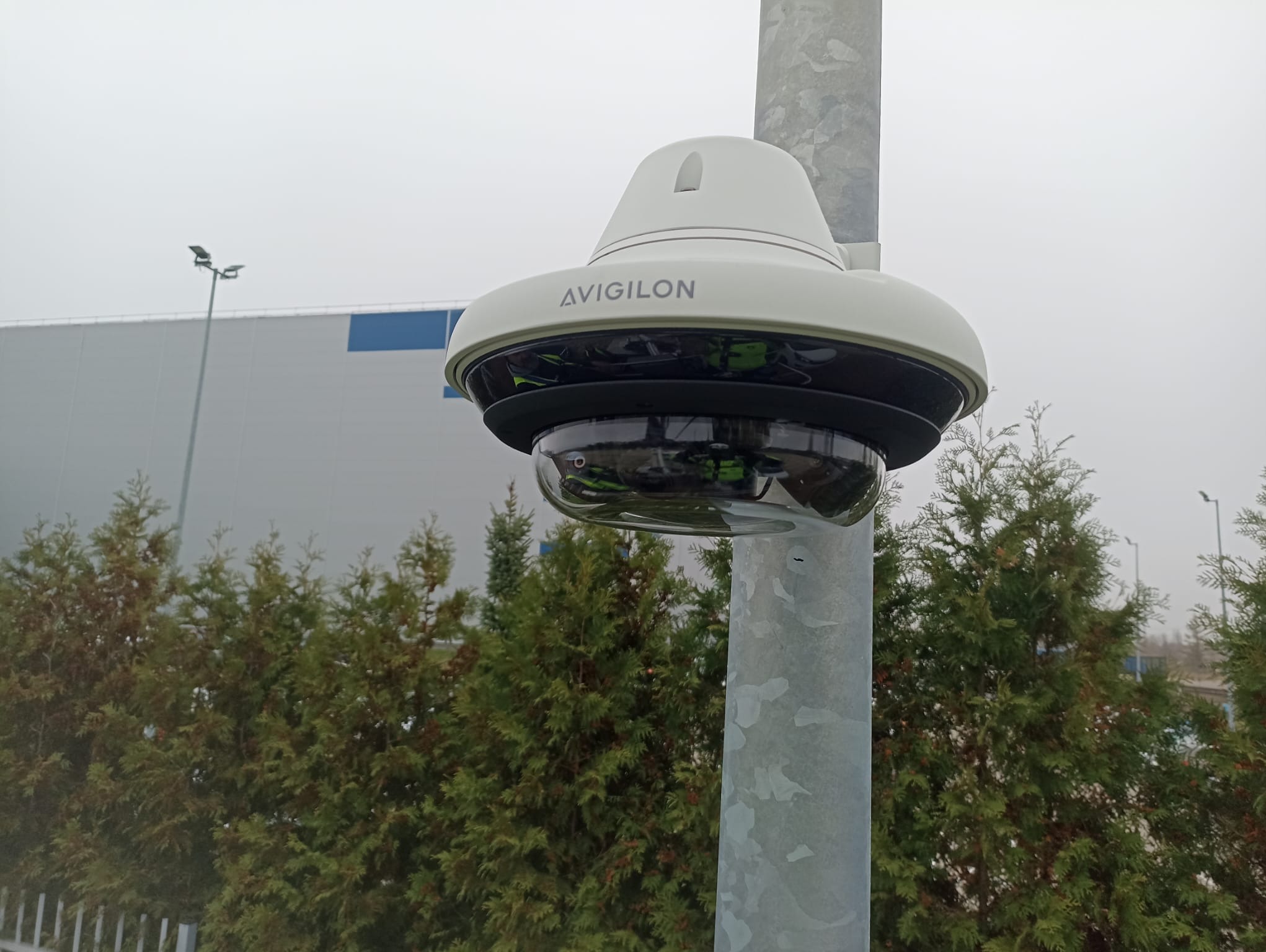 Kamera przemysłowa Avigilon zamontowana na metalowym słupie, na tle zielonych drzew i hali magazynowej w pochmurny dzień.
