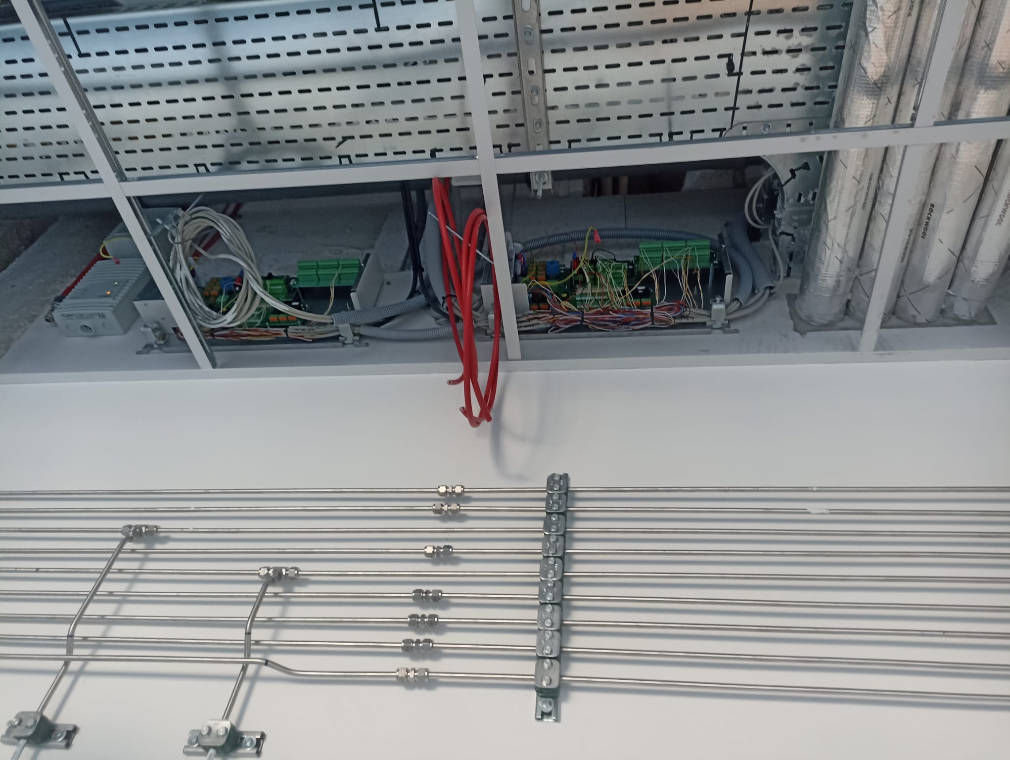 Widok instalacji elektrycznej i rur wentylacyjnych pod sufitem podwieszanym z widocznymi przewodami, puszkami i elementami mocującymi.