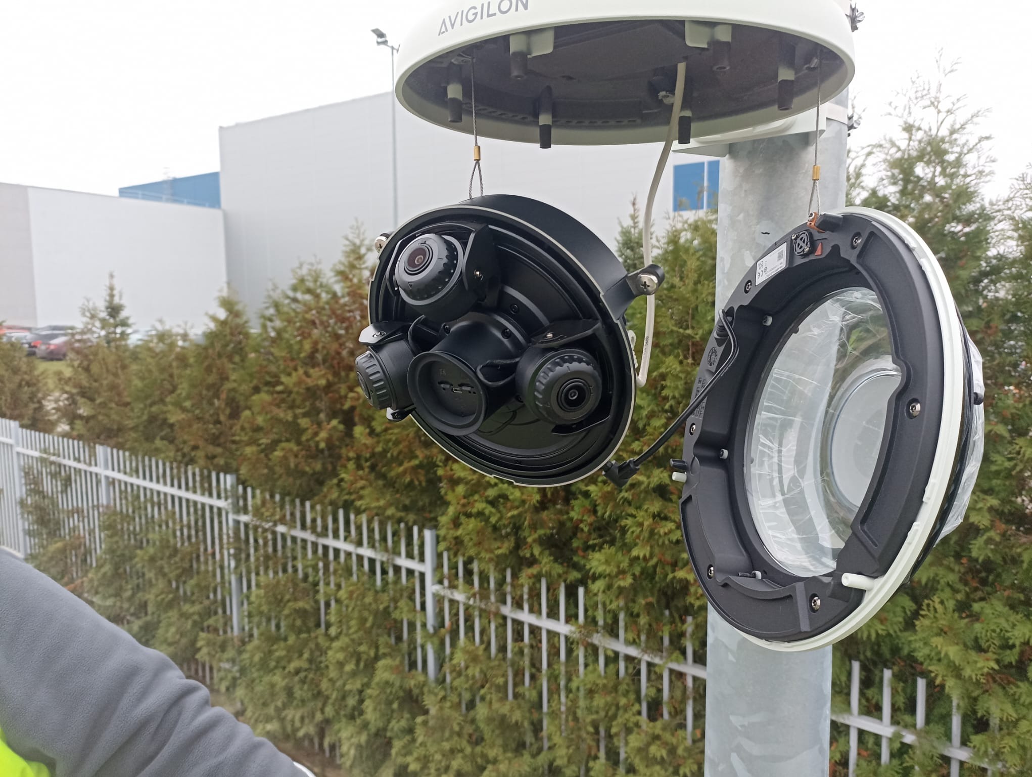 Otwarta obudowa kamery monitoringu Avigilon na słupie, widoczne kamery wewnątrz, tło z zielonymi krzewami i szarym budynkiem.