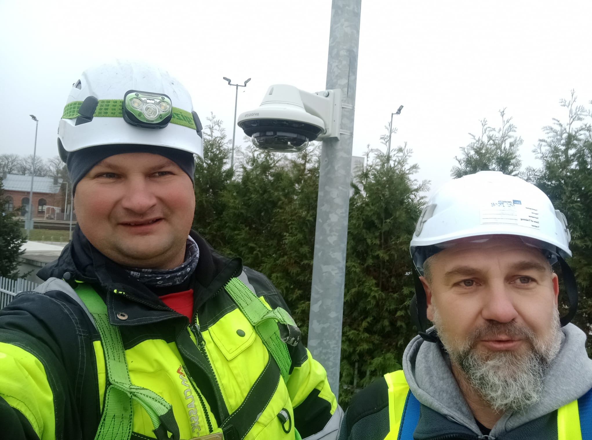 Dwóch mężczyzn w kaskach i kamizelkach odblaskowych pozuje przy słupie z kamerą monitoringu, widoczne drzewa w tle.