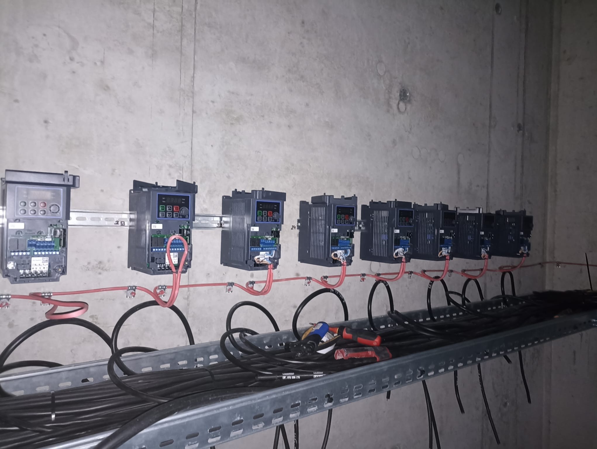 Szereg zamontowanych falowników na szynie DIN, z widocznymi przewodami i narzędziami na korytku kablowym, na tle surowej betonowej ściany.