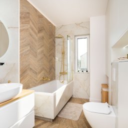 POD KLUCZ SPÓŁKA Z OGRANICZONĄ ODPOWIEDZIALNOŚCIĄ - Nowoczesna łazienka z wanną obudowaną marmurem, złotą armaturą, umywalką na drewnianym blacie i ścianą w jodełkę z płytek imitujących drewno.