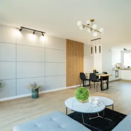 Jasne, przestronne wnętrze salonu z aneksem kuchennym, ściana z szarych paneli, drewniane lamele, parkiet ułożony w jodełkę, nowoczesne oświetlenie punktowe i wiszące, szara sofa i marmurowe...