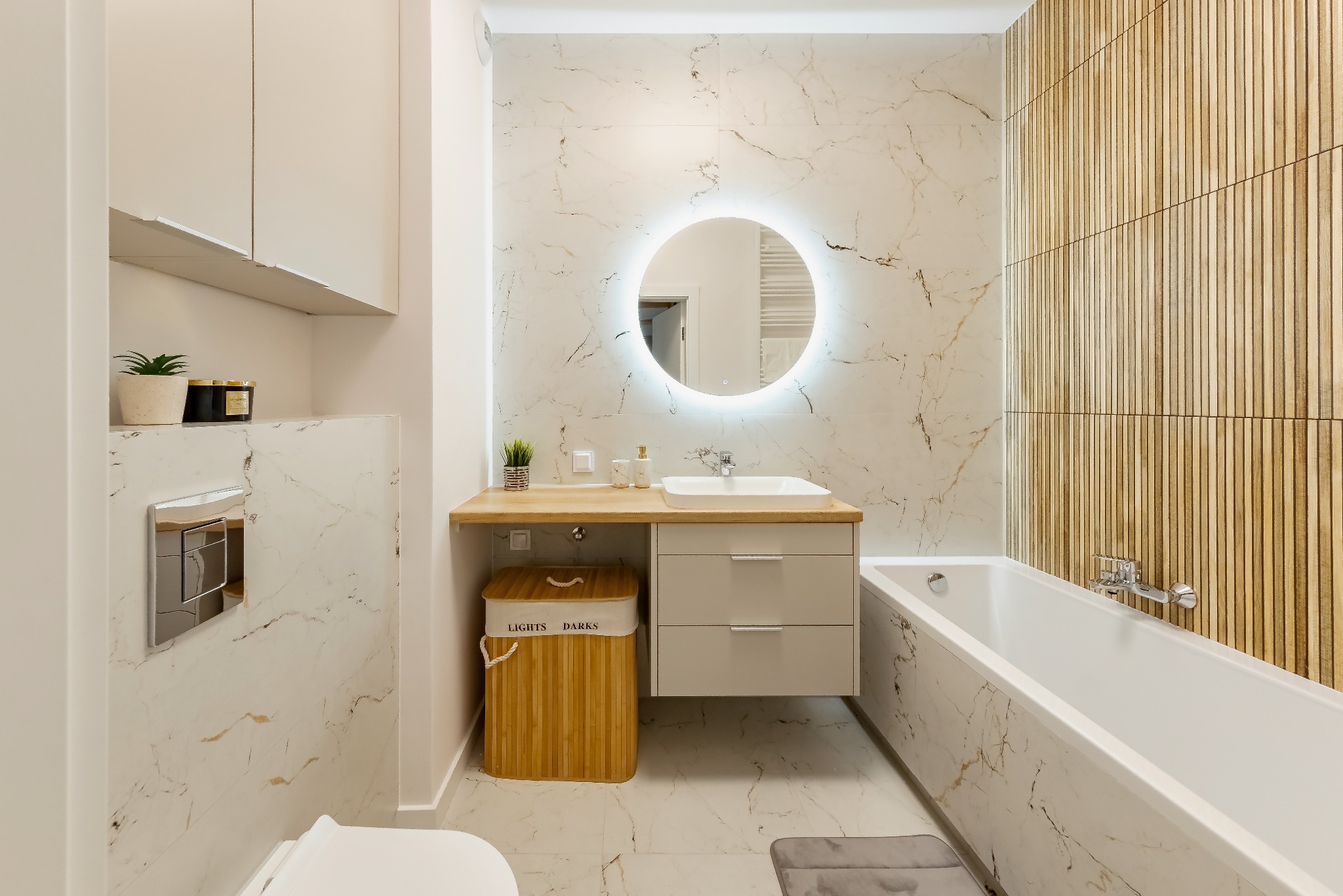 Nowoczesna łazienka z marmurowymi płytkami, okrągłym lustrem z podświetleniem LED, drewnianą ścianą i bambusowym koszem na pranie z napisem 'Lights Darks'.