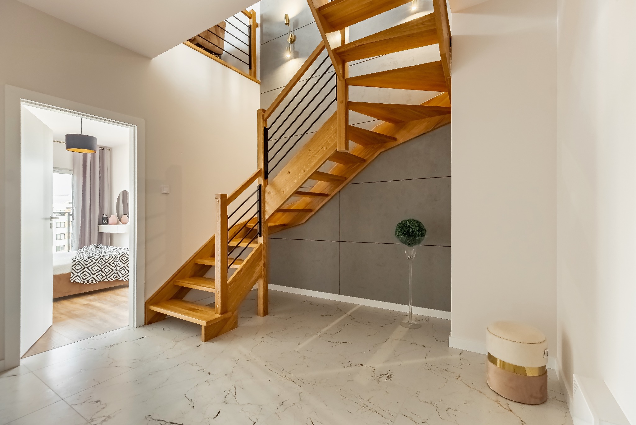 Jasne drewniane schody z czarnymi metalowymi barierkami w nowoczesnym wnętrzu z marmurową podłogą i szarą ścianą akcentującą, widok na otwarty pokój z łóżkiem.