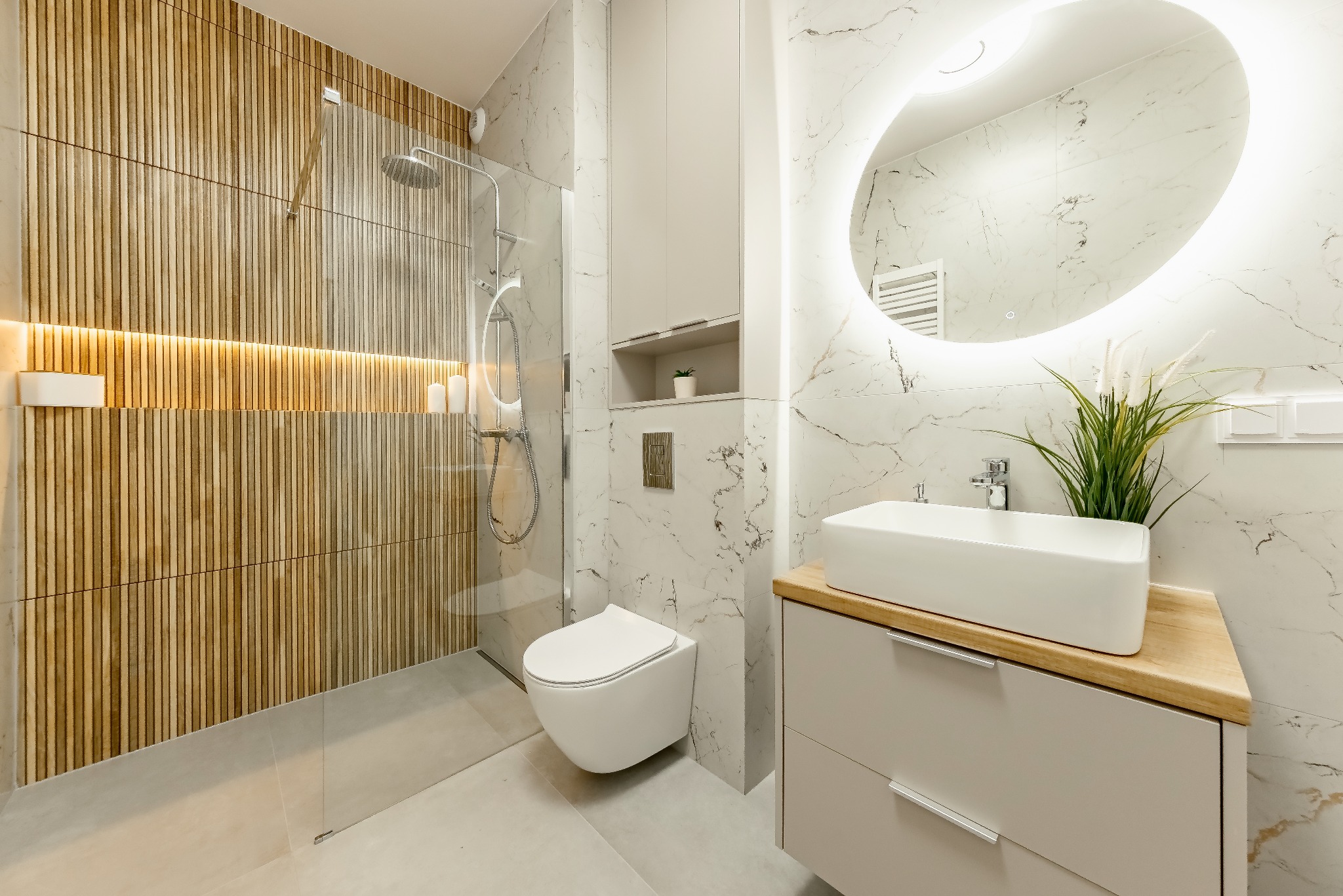 Nowoczesna łazienka z prysznicem typu walk-in, umywalką nablatową na drewnianym blacie i podświetlanym lustrem owalnym, wykończona płytkami imitującymi marmur i drewno.