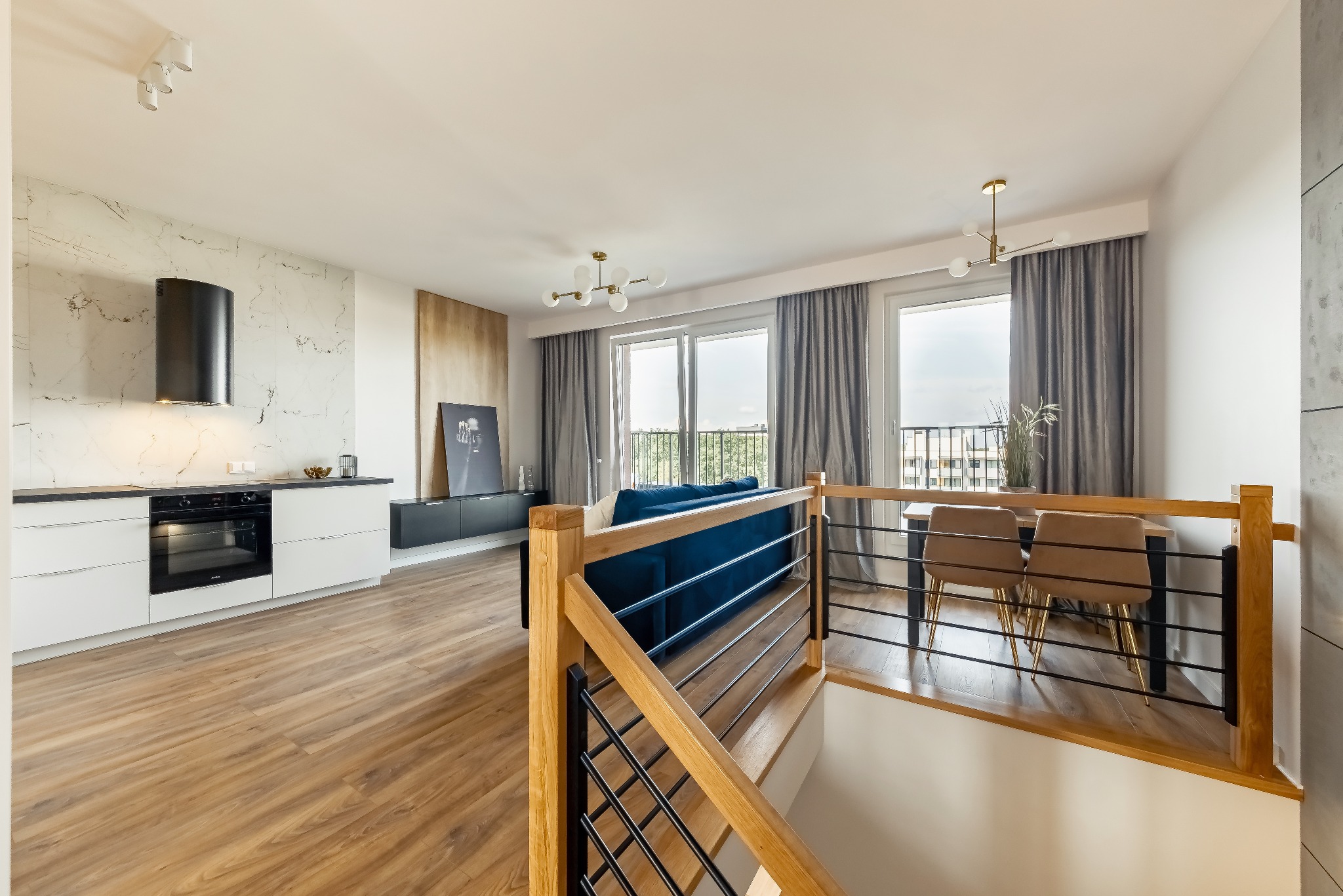 Nowoczesne wnętrze domu z drewnianą podłogą, białą kuchnią z czarnym okapem, marmurową ścianą, schodami z drewnianą balustradą i salonem z niebieską sofą w tle.