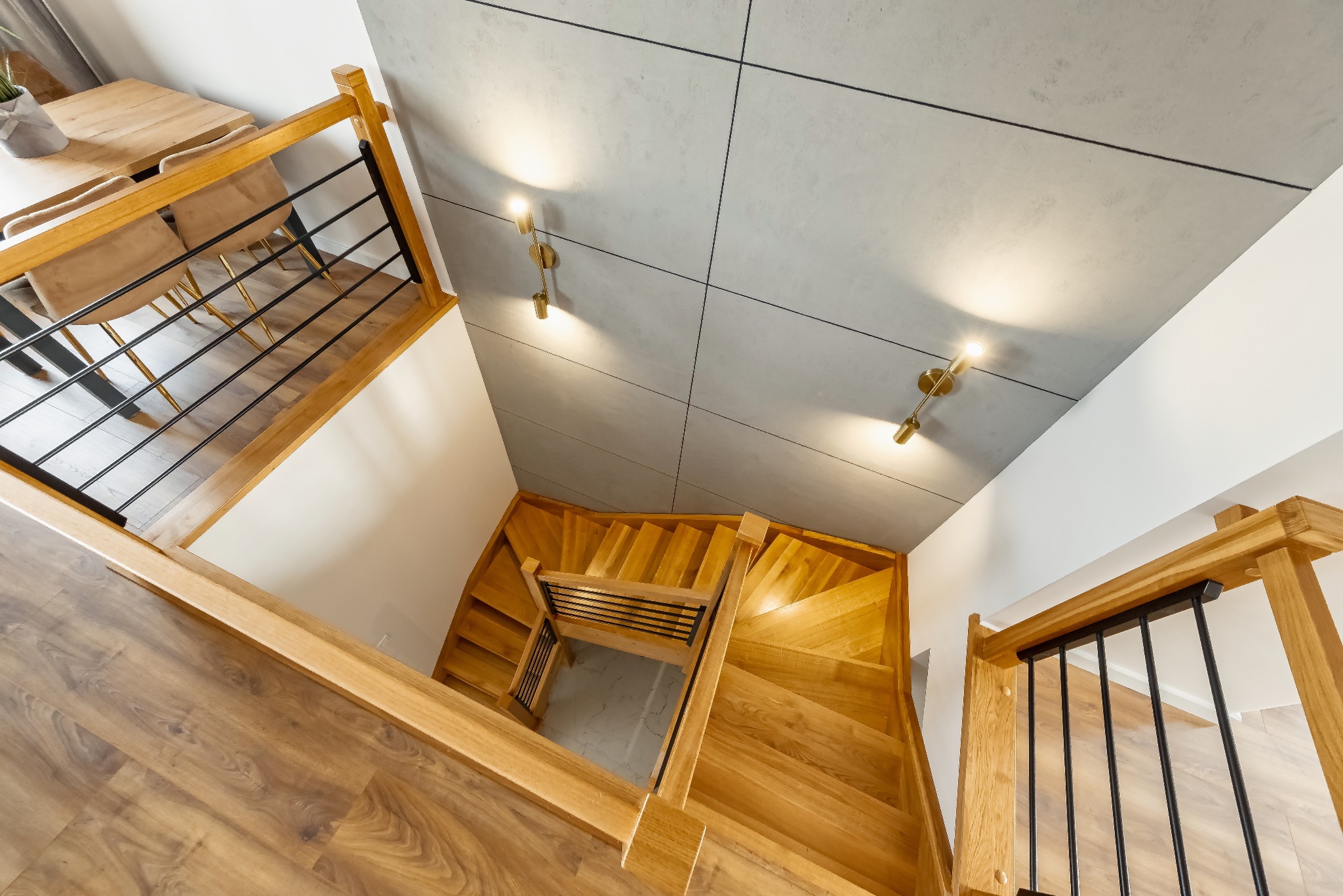 Widok z góry na drewniane schody z czarnymi metalowymi balustradami, szare płytki na suficie z nowoczesnymi złotymi lampami ściennymi, fragment drewnianego stołu i krzeseł w tle.