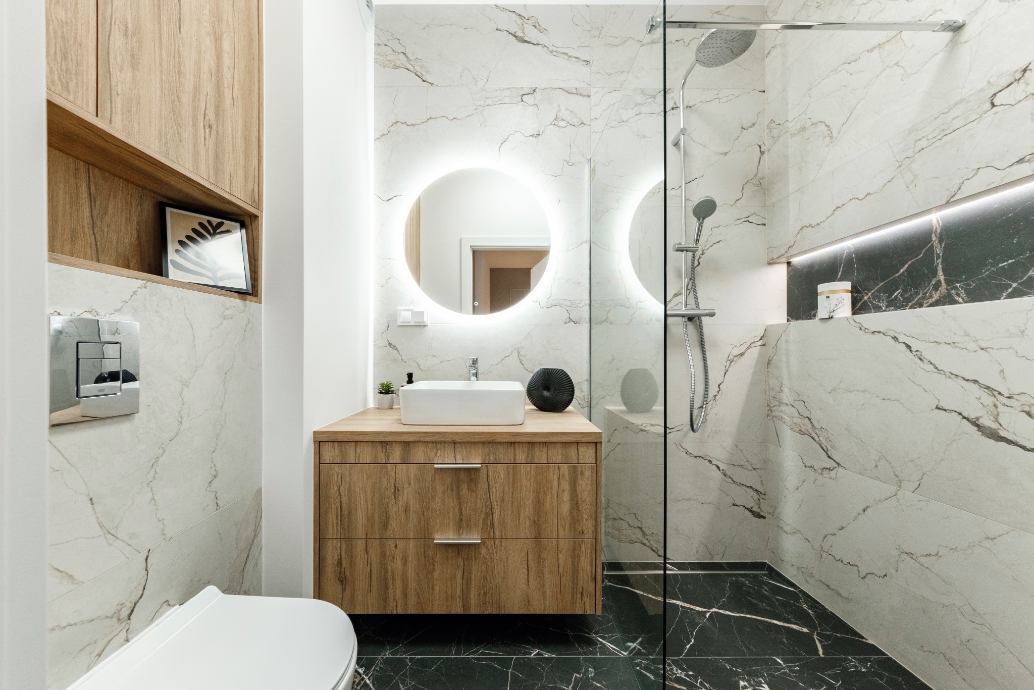 Nowoczesna łazienka z marmurowymi płytkami, okrągłym lustrem z podświetleniem LED, drewnianą szafką pod umywalką i czarną podłogą z marmuru.