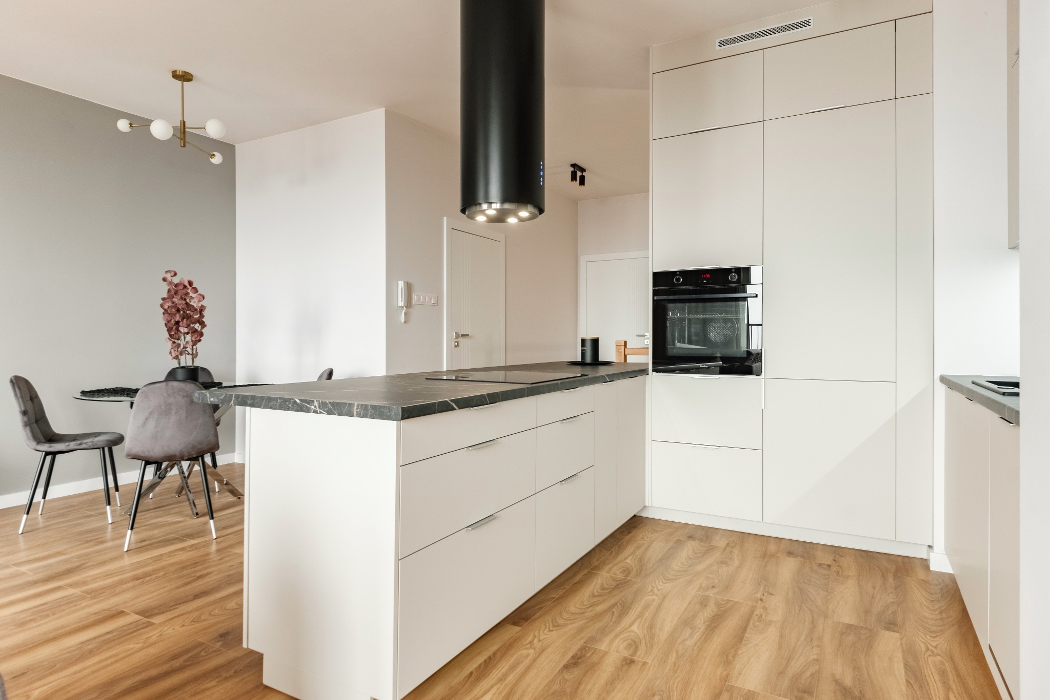 Nowoczesna, jasna kuchnia z wyspą, czarnym okapem cylindrycznym i drewnianą podłogą, widok na jadalnię z szarymi krzesłami.