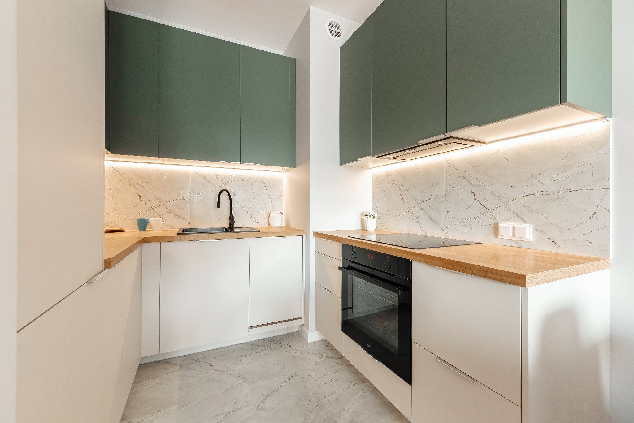 Nowoczesna kuchnia w bieli i zieleni z drewnianym blatem, marmurową ścianą i czarną armaturą, oświetlona ledowym światłem pod szafkami.