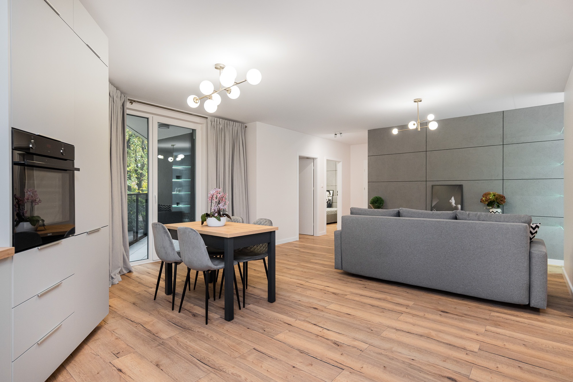 Jasne, przestronne wnętrze salonu z drewnianą podłogą, szarym narożnikiem, stołem z kwiatami i nowoczesnym oświetleniem.