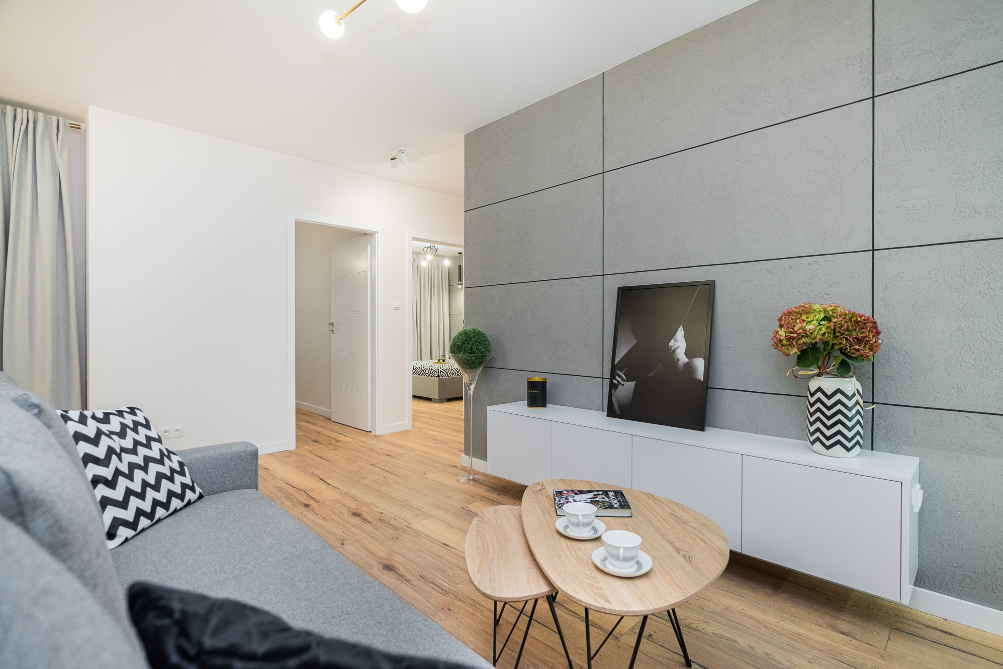 Nowoczesny salon z szarą ścianą z paneli, drewnianą podłogą, szarą kanapą z wzorzystymi poduszkami, białymi meblami i stolikami kawowymi z filiżankami.